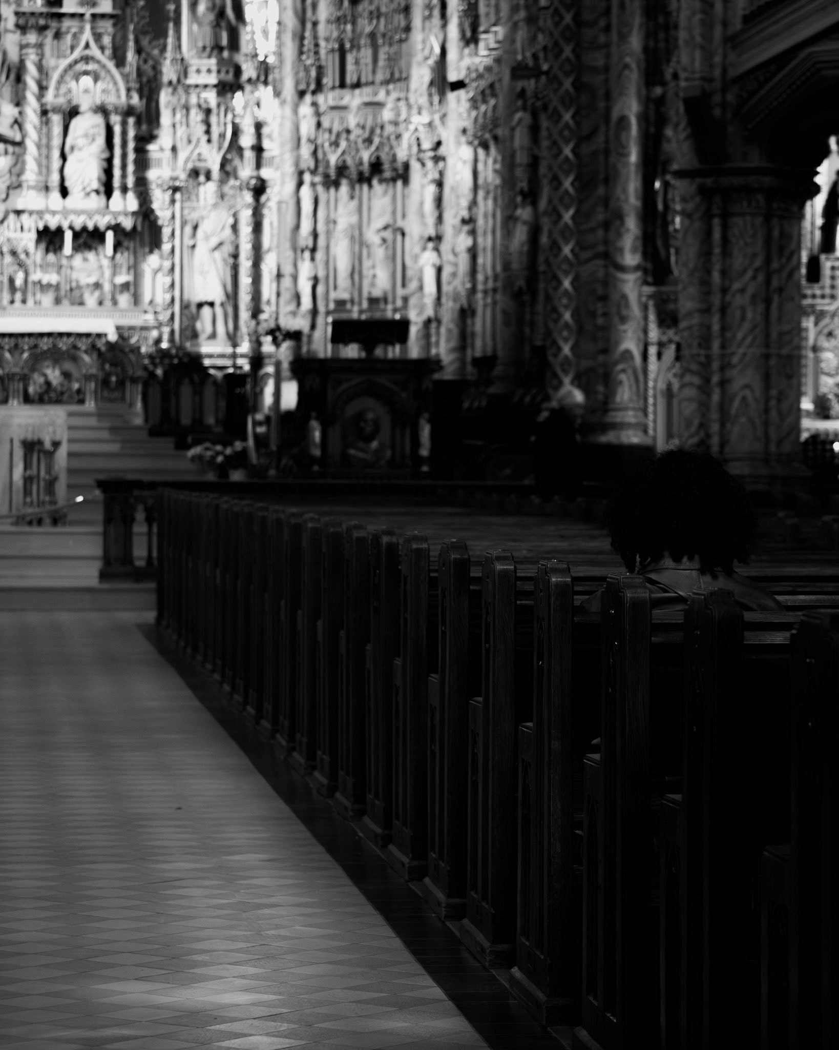 Lady in a Church: A monochrome interior shot looking down the long, tiled aisle of a grand, ornate church. In the foreground, a woman sits alone in a wooden pew, her back to the camera, captured in soft shadow. The background shows the intricate, carved details of the altar and pillars, bathed in soft, diffused light, creating a deep sense of depth and architectural reverence.