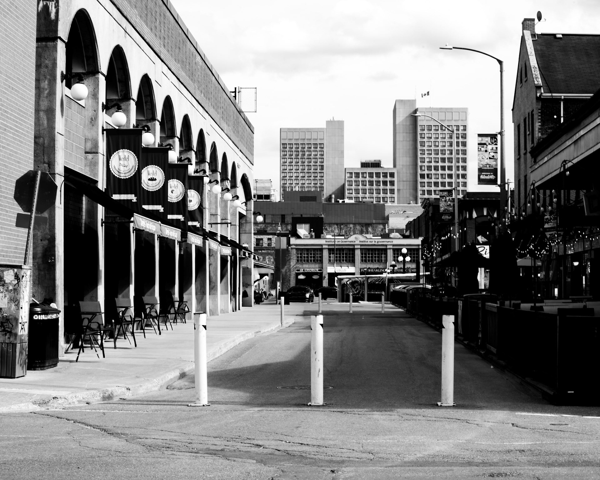Empty Arcade: Monochrome fine art street photography of a deserted urban street lined with arched stone porticos, heavy banners, and bollards leading toward distant downtown high-rises.