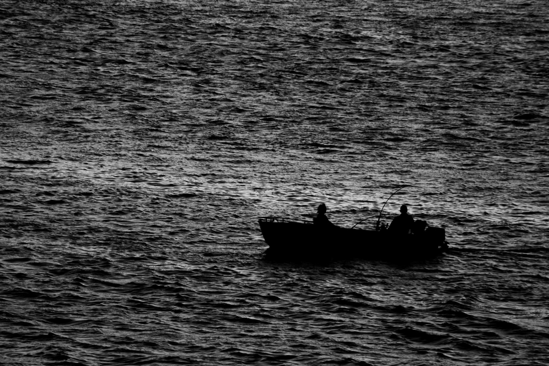 Communion: Monochrome fine art photography capturing two silhouetted figures in a small boat floating on a vast, textured expanse of dark water under a moody sky.