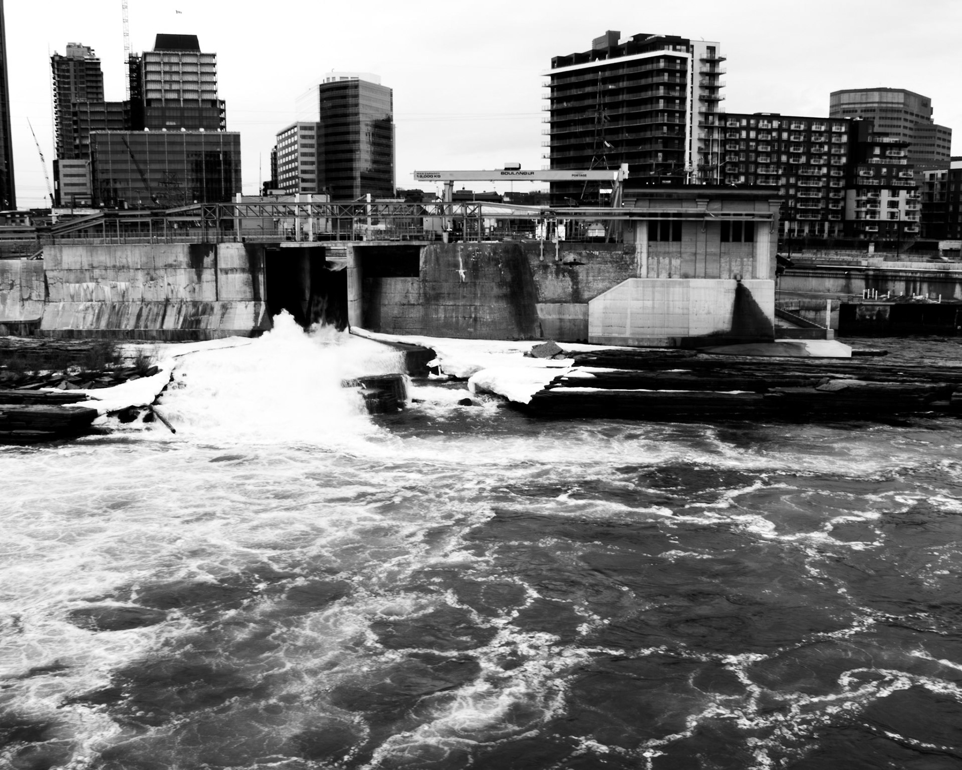 Structured Waters: Monochrome fine art architectural photography capturing a powerful industrial dam and churning whitewater release, set against a backdrop of stark urban high-rises and geometric concrete infrastructure.