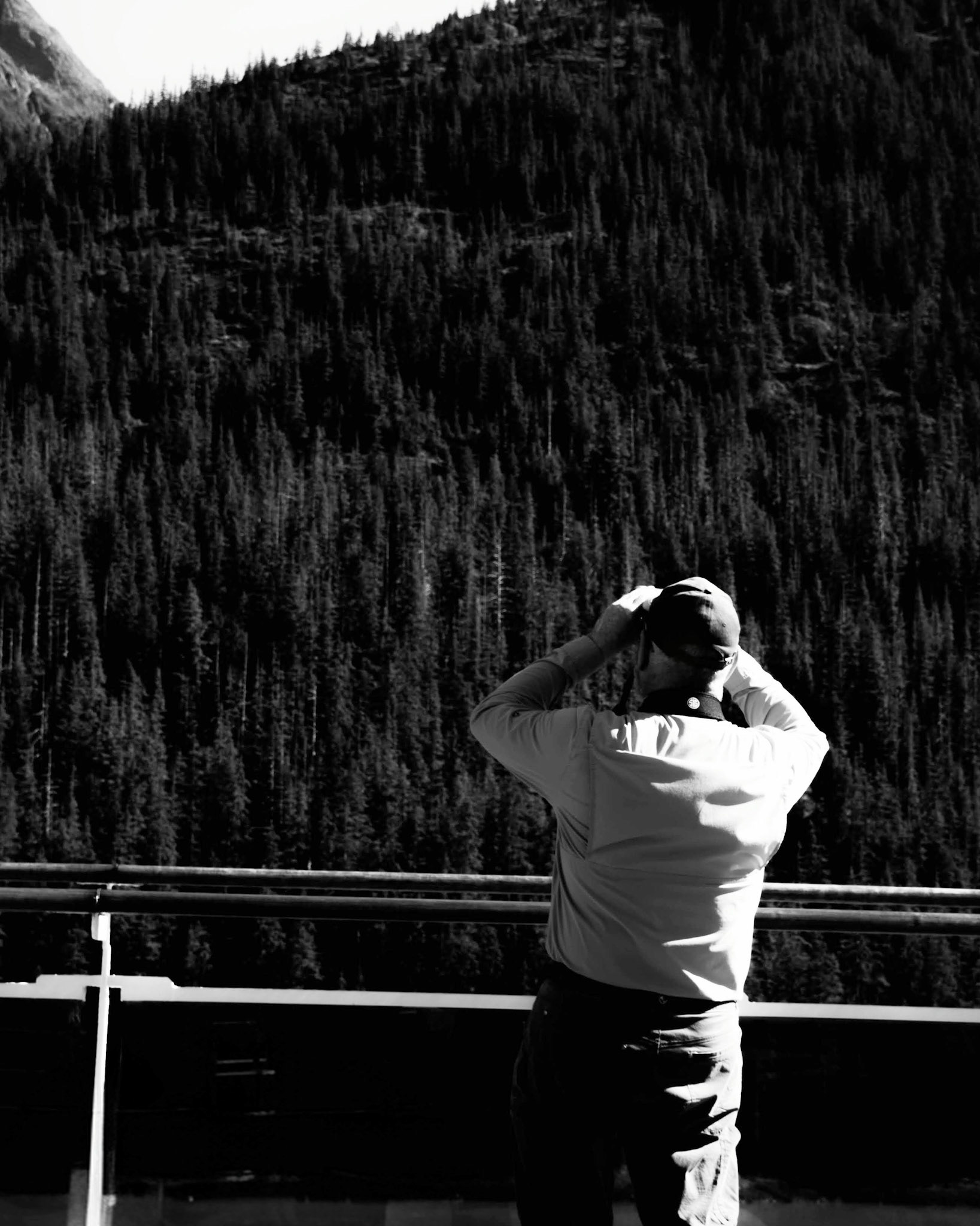 The Scholar: A monochrome photograph from behind a man standing at a clean, white railing. He is holding binoculars to his eyes, looking intently toward a vast, dense forest of evergreen trees that fills the entire background. The strong contrast between the bright railing and the dark, textured forest highlights the subject’s focus and the scale of the natural landscape.