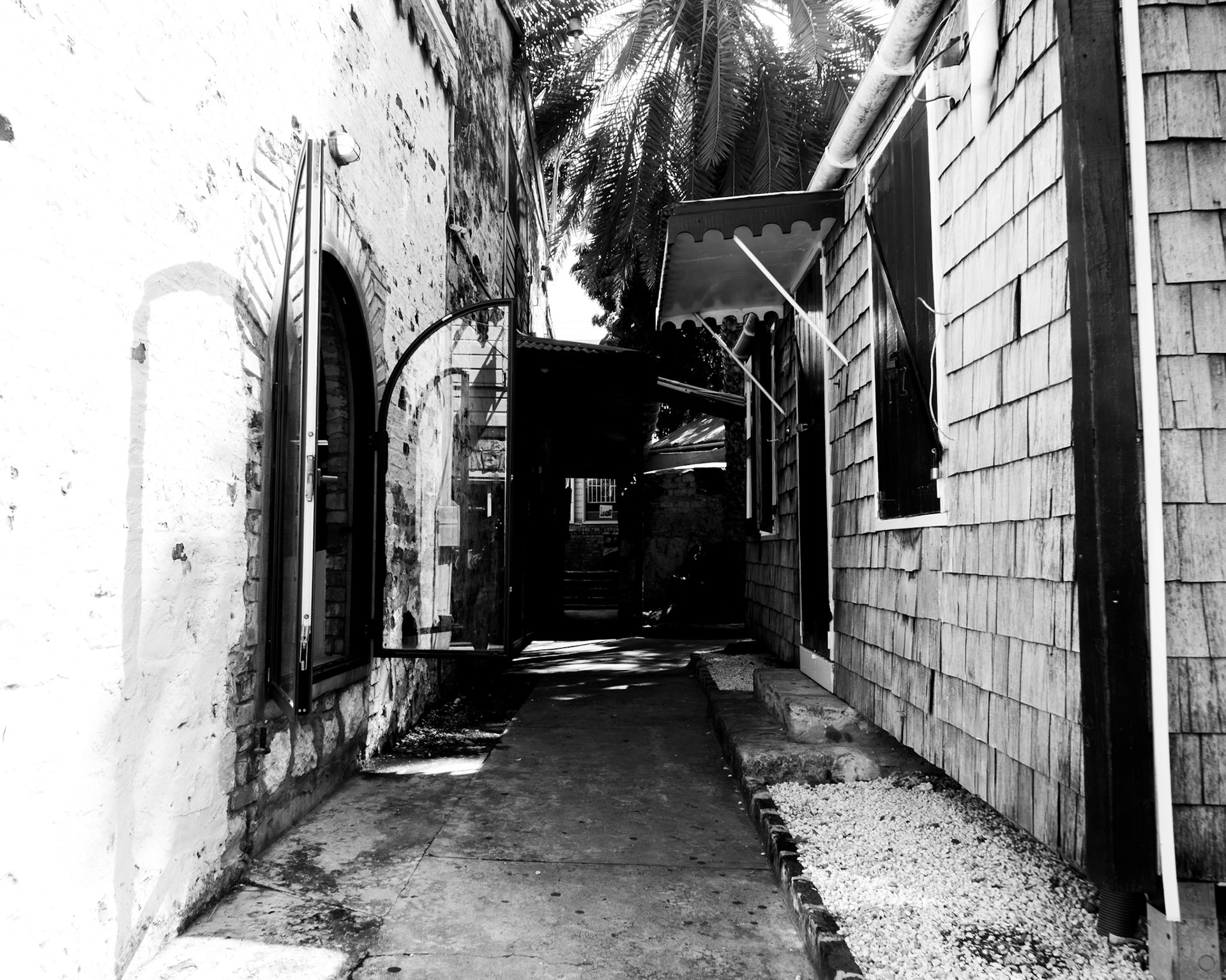 Alley of Light: Monochrome fine art architectural photography capturing a narrow, sun-drenched passageway between textured historic buildings, framed by open architectural glass and deep, contrasting shadows.