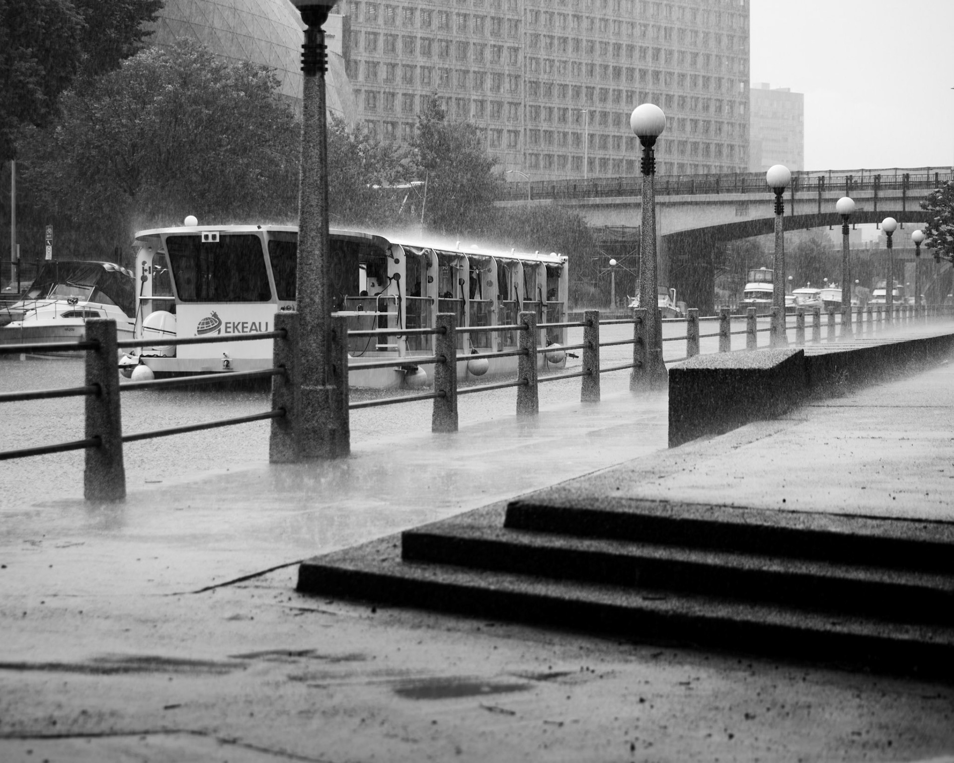 Canal of Rain: Monochrome fine art photography capturing a heavy downpour over a deserted urban canal walkway, featuring rain-streaked concrete steps, vintage globe lamps, and a lone tour boat moored against the railing.
