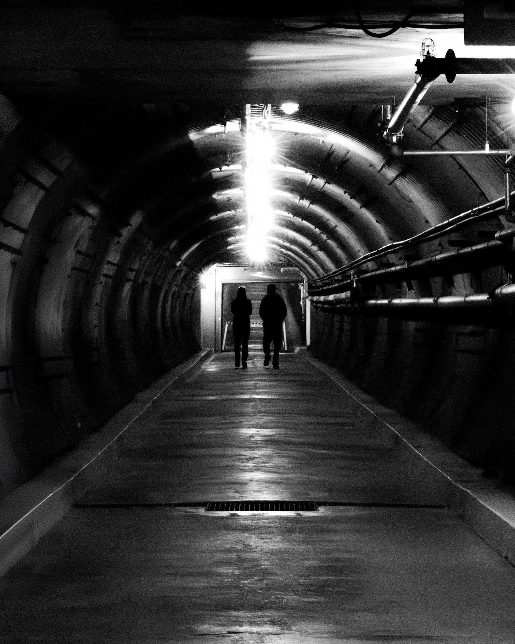 The Pilgrimage: A monochrome, symmetrical photograph looking down a long, curved industrial tunnel. Two figures are silhouetted in the distance, walking toward a bright light source at the far end of the passage. The composition is defined by the dark, textured, ribbed walls of the tunnel, overhead pipes, and strong leading lines that draw your eyes toward the subjects and the brilliant light.