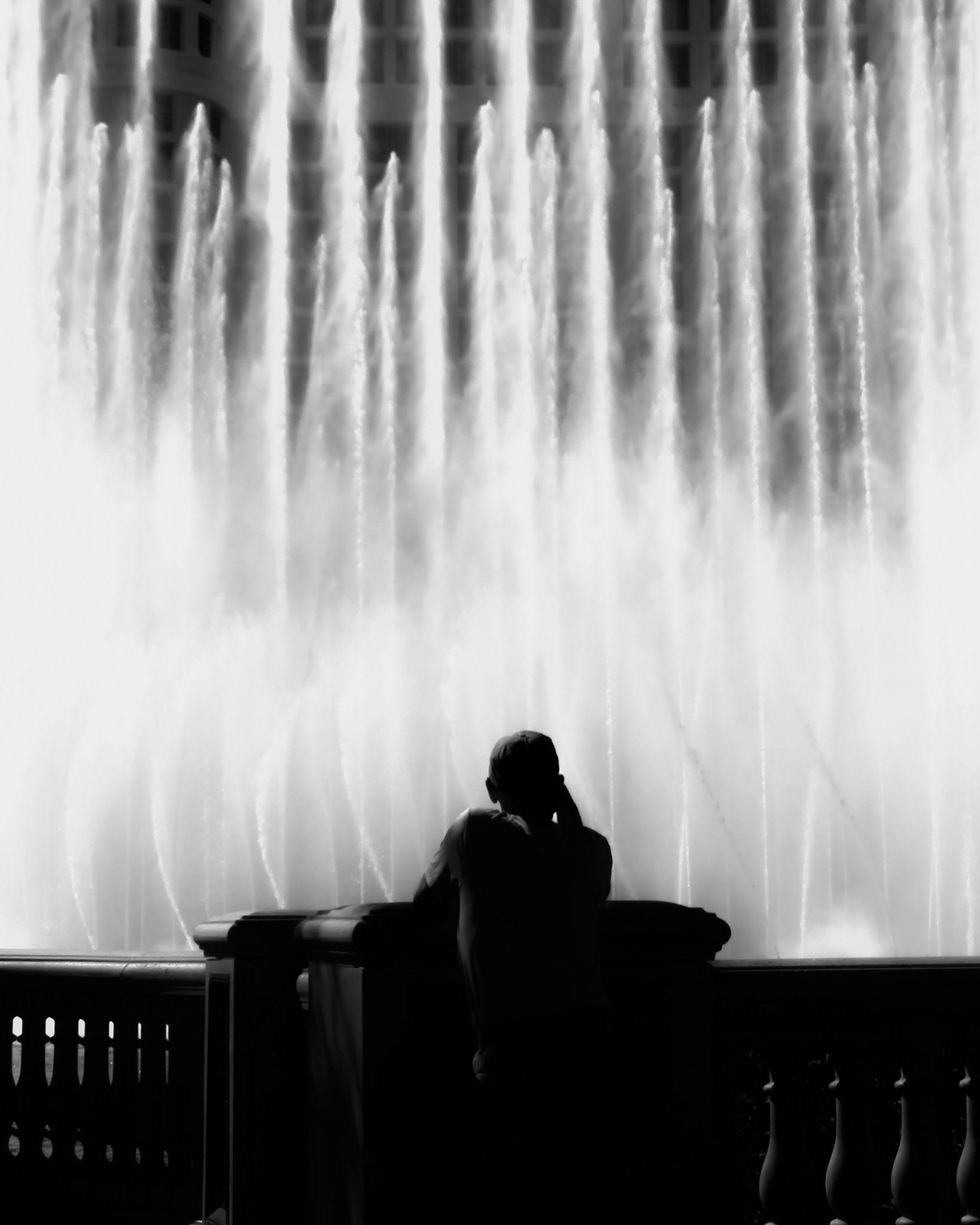 Liquid Alter: A monochrome photograph showing the silhouette of a person leaning against a stone railing, facing a massive, glowing fountain. The water shoots upward in vertical, ethereal streams that blur into bright white light, creating an effect reminiscent of sunlight streaming through stained glass.