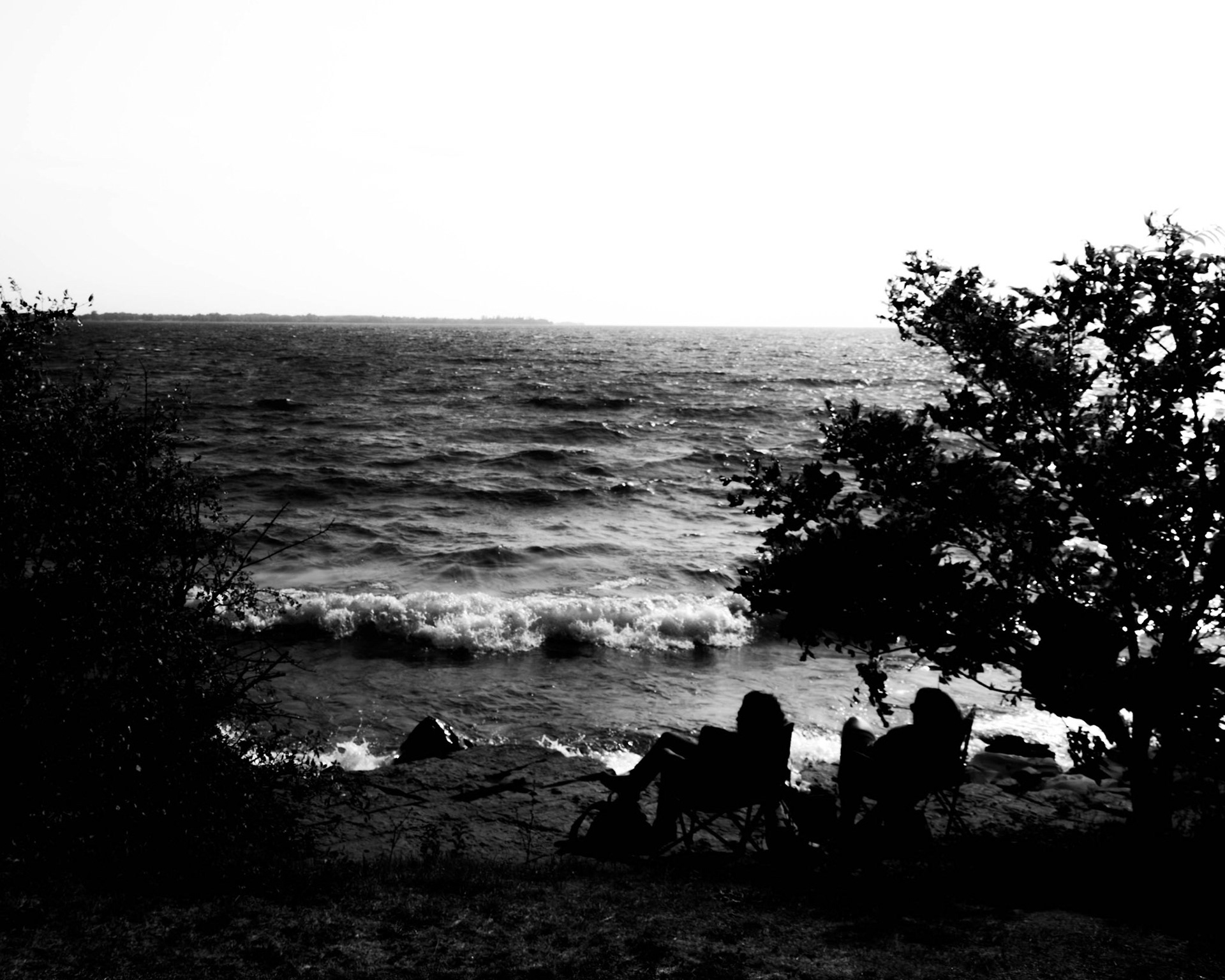 Cathedral: Monochrome fine art photography of two figures relaxing in lawn chairs on a rocky shoreline beneath framing trees, looking out over a vast, textured expanse of dark water under an open sky.