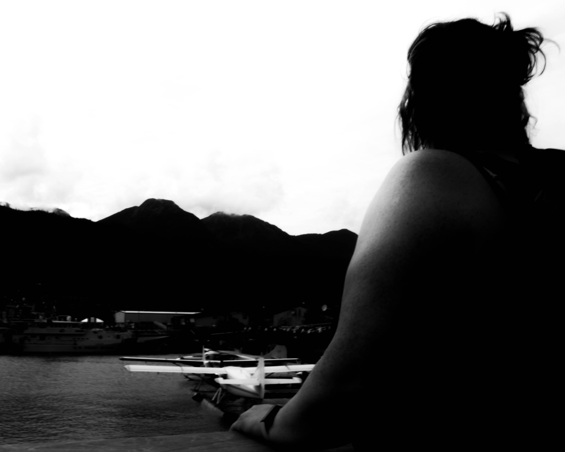 Traveller: Monochrome fine art photography from a low vantage point featuring a dark silhouette leaning against a railing overlooking a harbor, with a small seaplane and distant mountains under a bright sky.