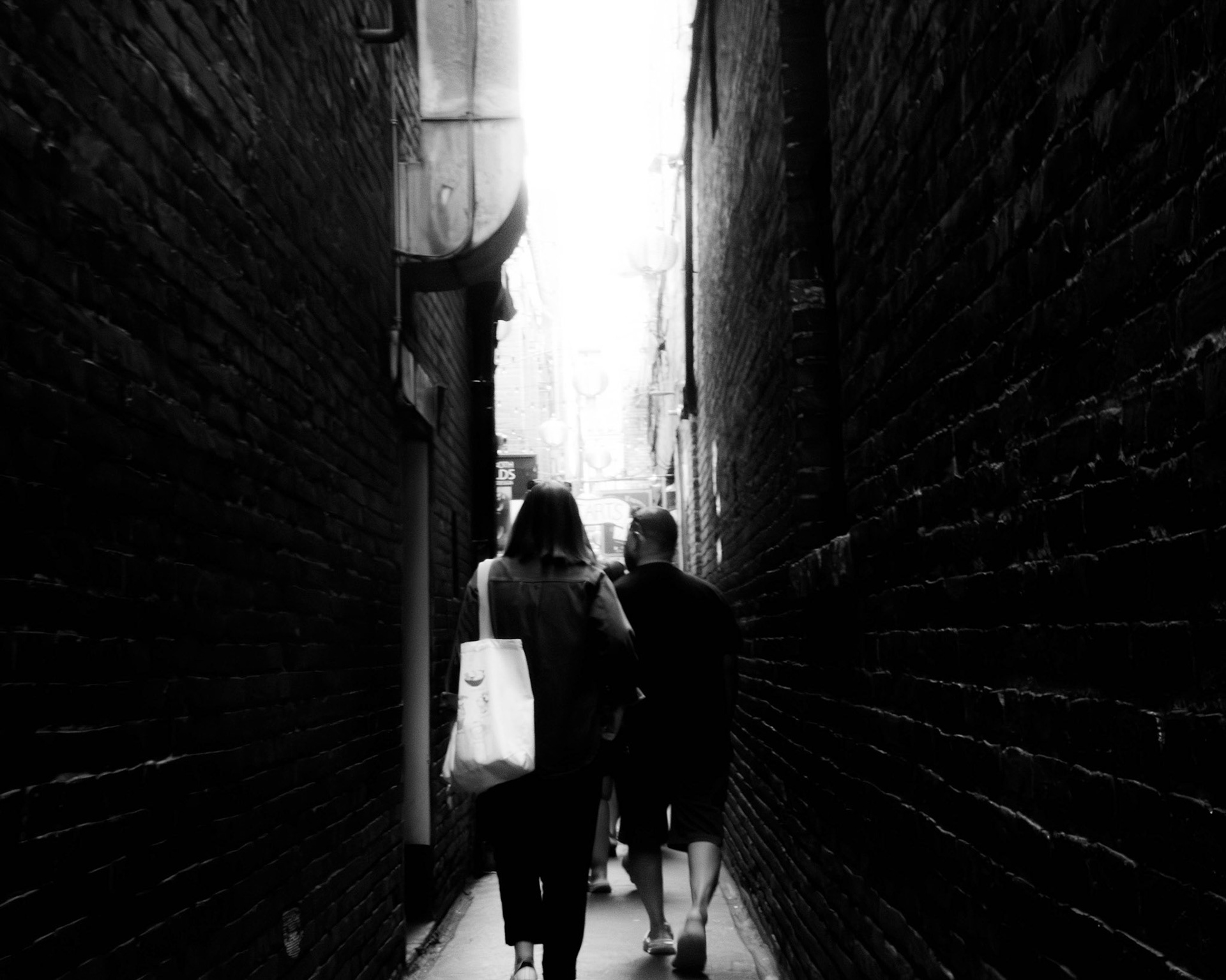 Alleyway Lovers: A monochrome photograph taken from behind a couple as they walk away from the viewer through a narrow, dark brick alleyway. Their figures are silhouetted against a brilliant, blown-out light source at the end of the passage, emphasizing the texture of the brick walls and the quiet solitude of the scene..