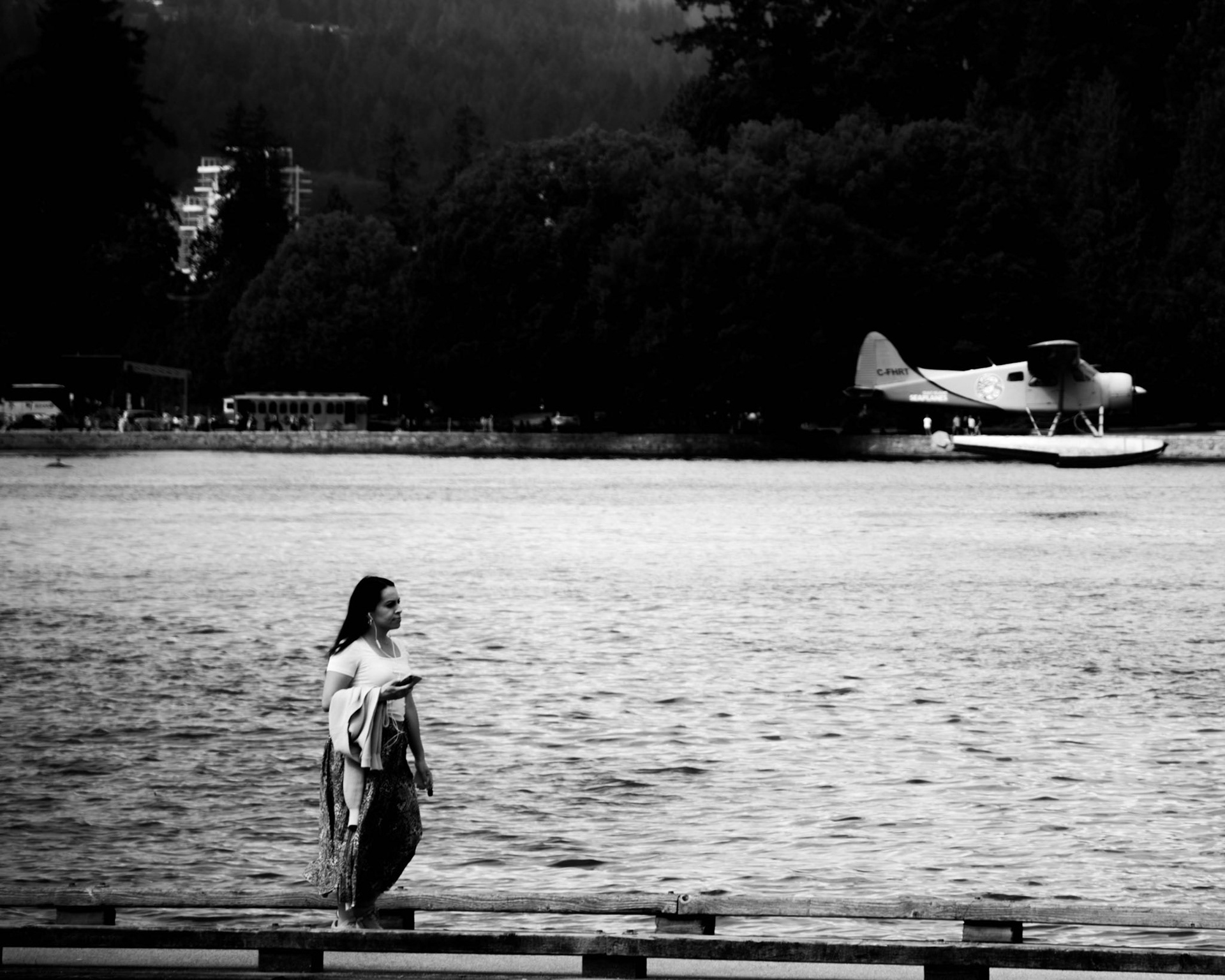 Hark, Ye Angel: Monochrome fine art street photography capturing a lone woman walking along a wooden boardwalk beside dark water, with a vintage seaplane hovering in the distance under a moody, overcast sky.