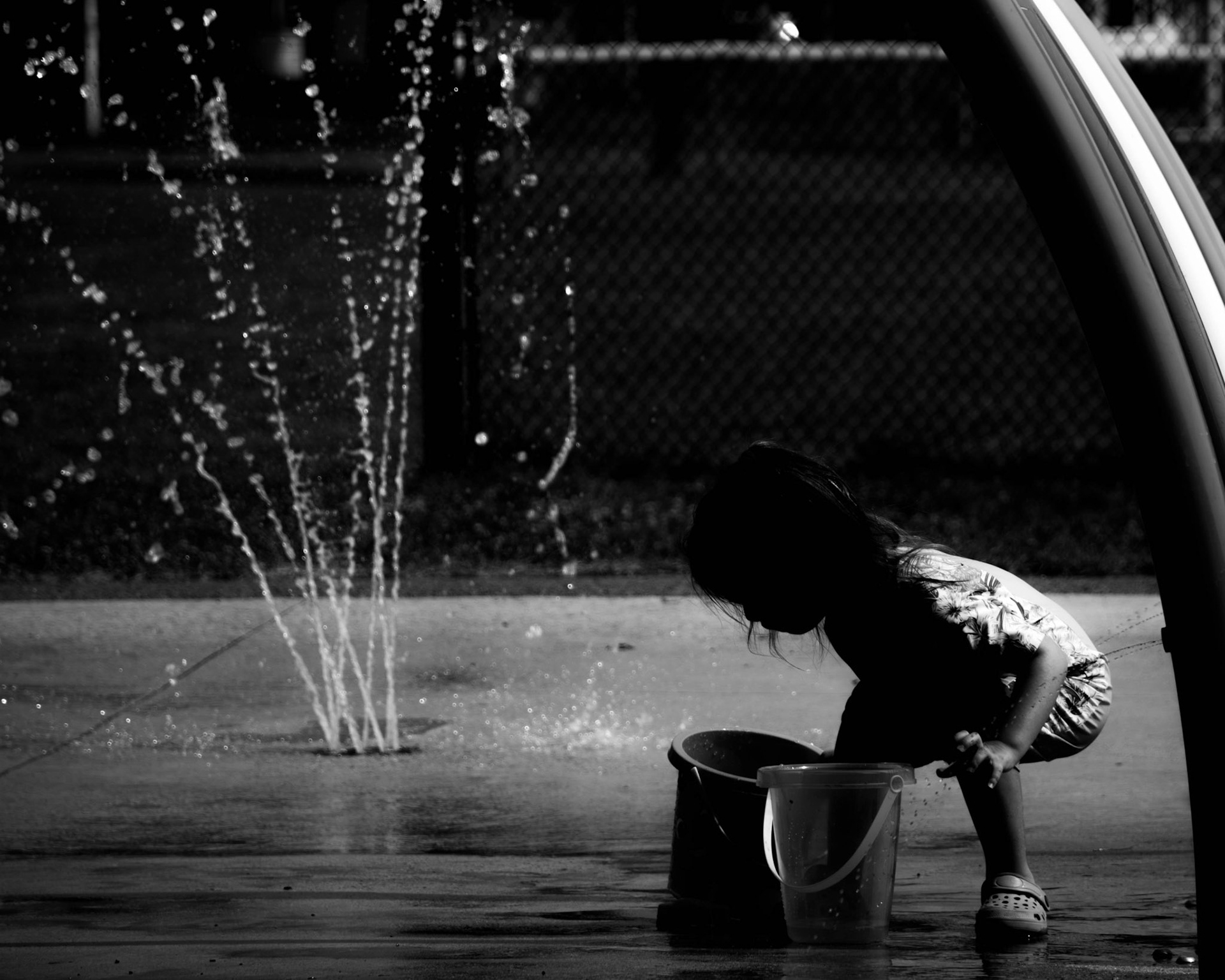 Holy Water: A monochrome photograph of a child crouching at a splash pad. The child is captured as a silhouette, intently focused on filling a plastic bucket with water from a nearby spray fountain. The scene is framed by a curved metal structure, and the contrast between the dark shadows and the sparkling water droplets creates a serene, ritualistic atmosphere.