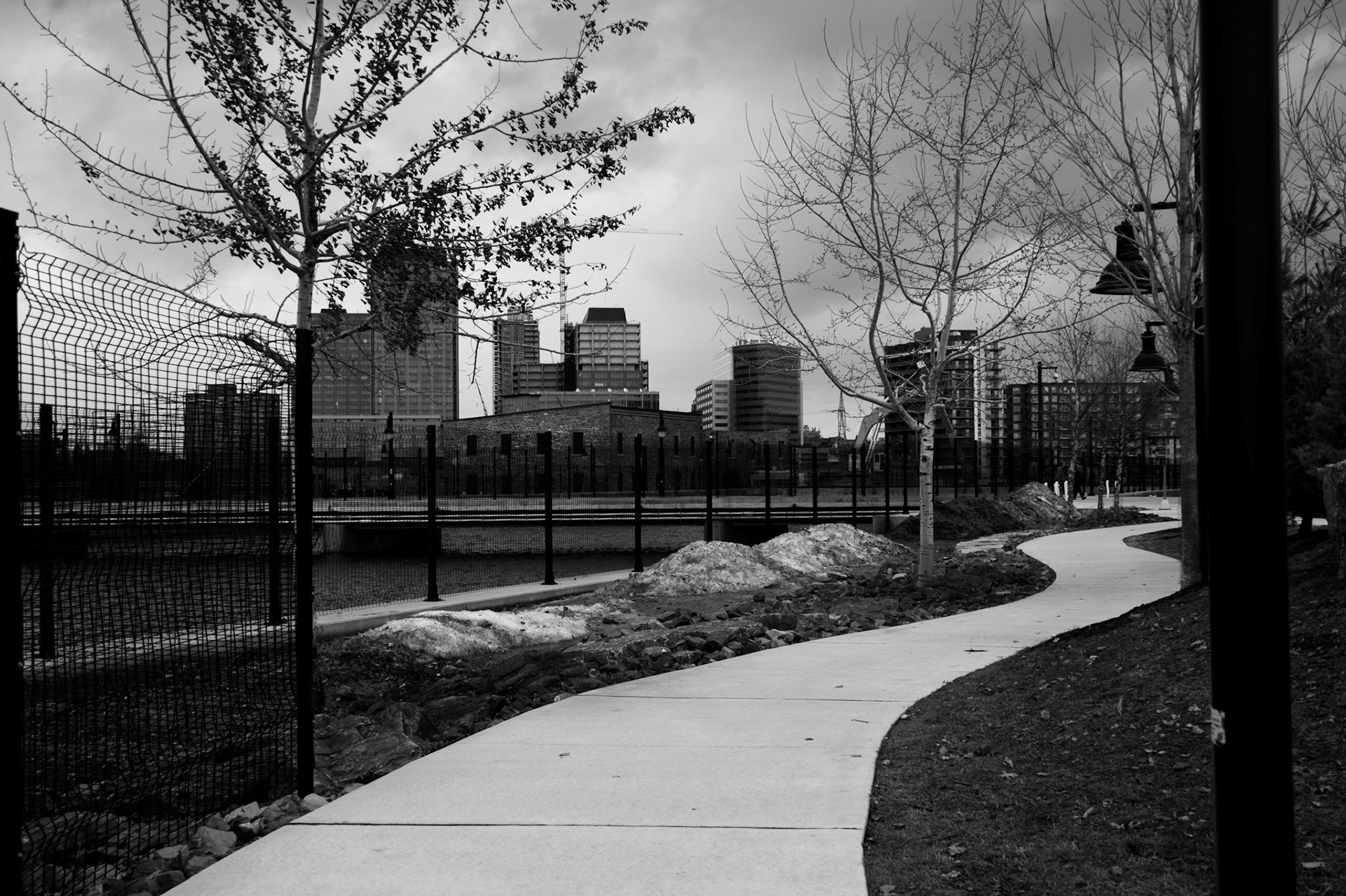 Structural Approach: Monochrome fine art photography capturing the geometric lines of an isolated concrete walkway as it approaches the urban core, defined by stark high-contrast light and shadow.