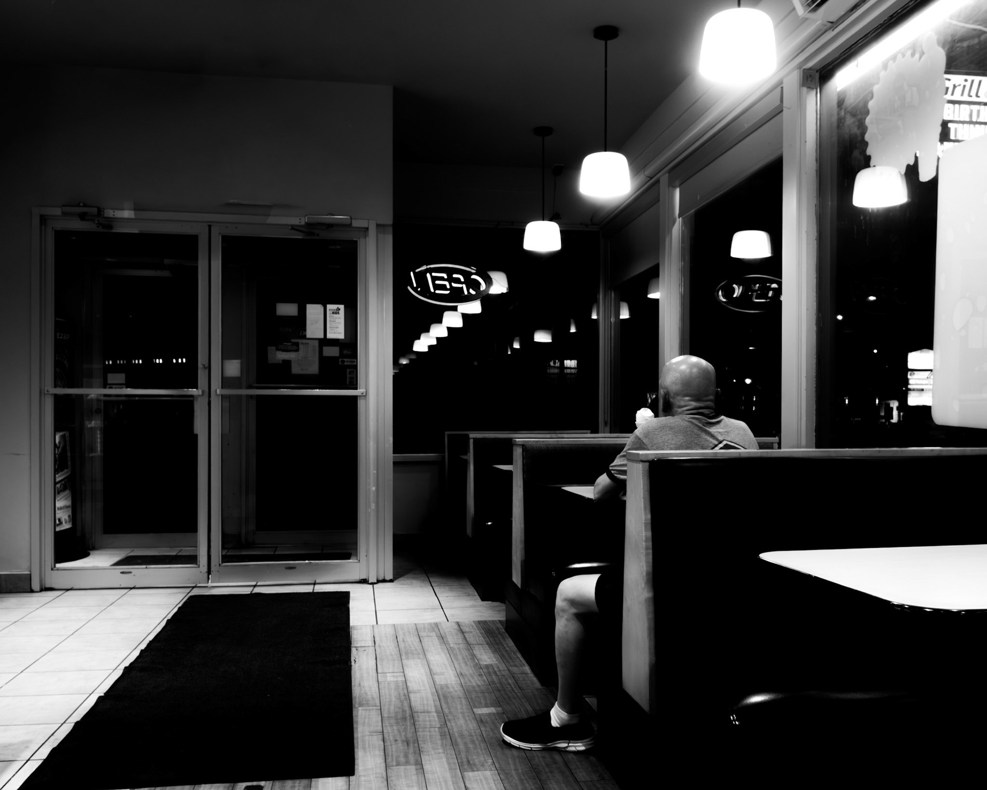 Man in a Diner: Monochrome interior fine art photography capturing a solitary figure seated in a booth, framed by high-contrast neon reflections and glass doors in an Ottawa eatery.