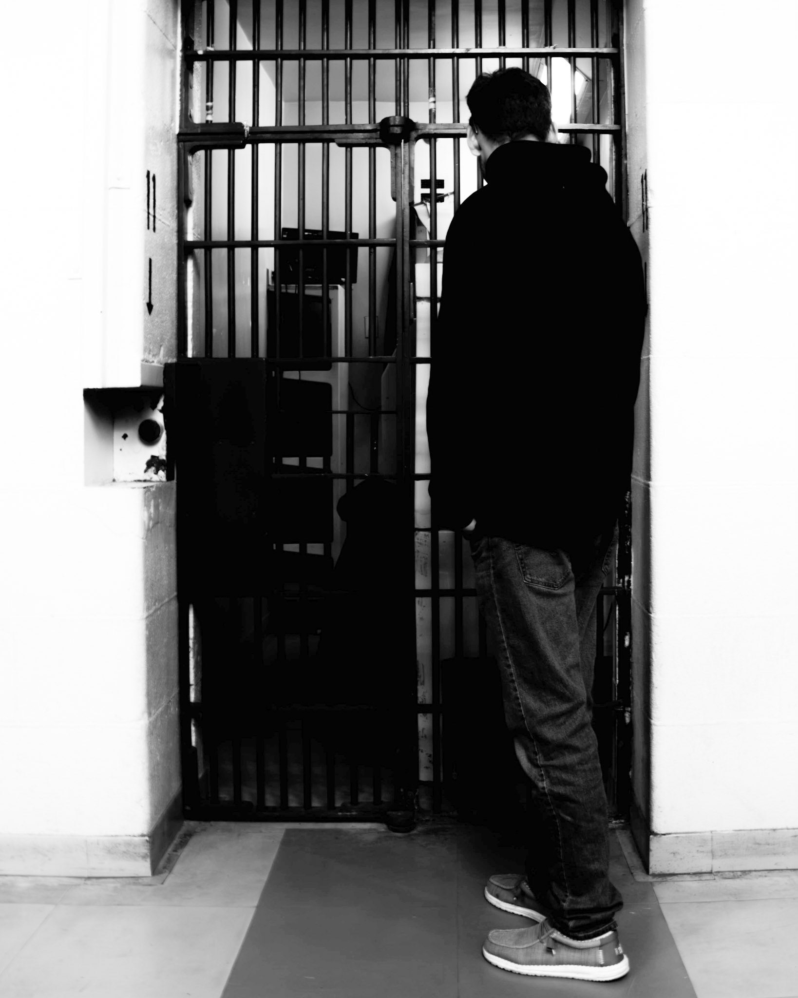 The Confessional: A monochrome photograph of a man standing in profile before a prison cell. The subject is captured in deep shadow, facing the dark interior behind the bars. The stark, high-contrast lighting emphasizes the geometric rhythm of the metal bars against the bright, plain white walls, creating a sense of confinement and introspective stillness.