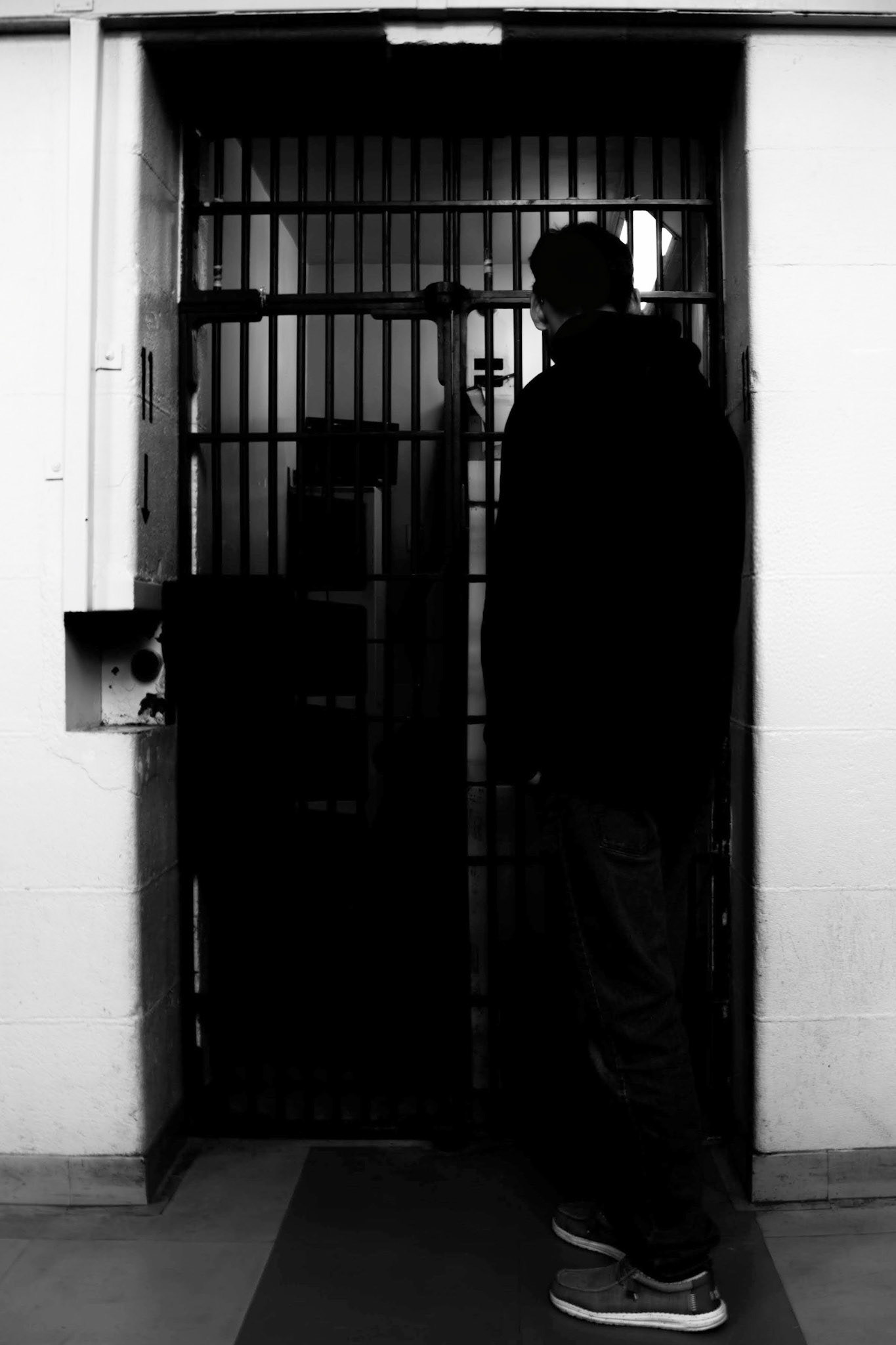 The Confessional: A monochrome photograph of a man standing in profile before a prison cell. The subject is captured in deep shadow, facing the dark interior behind the bars. The stark, high-contrast lighting emphasizes the geometric rhythm of the metal bars against the bright, plain white walls, creating a sense of confinement and introspective stillness.