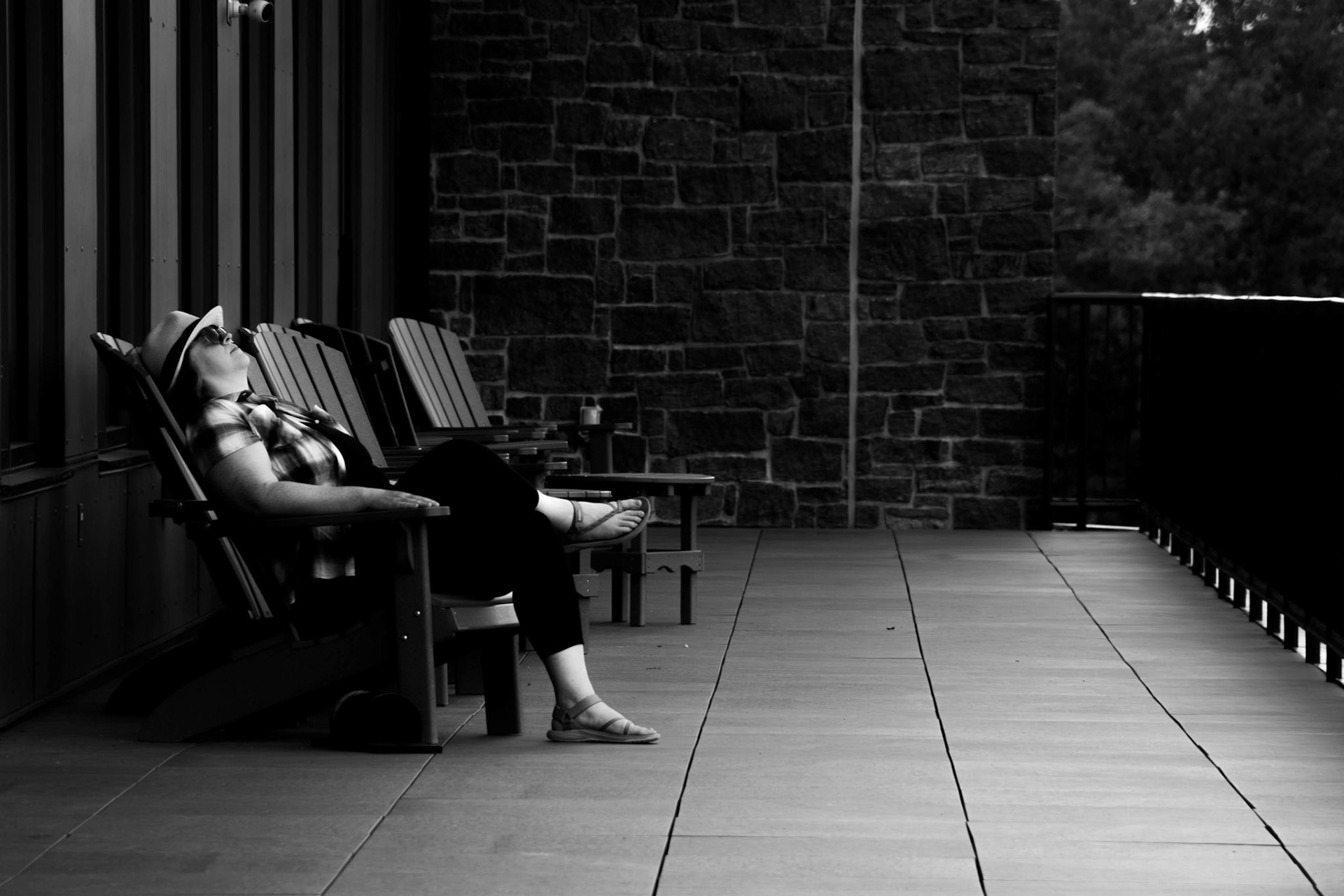 Meditation: A monochrome photograph of a woman resting in an Adirondack chair on a spacious balcony. The subject, wearing a hat and light clothing, is reclined with their eyes closed in a moment of stillness. The composition is defined by the strong, leading perspective lines of the floor tiles and railing, set against the backdrop of a textured stone wall.