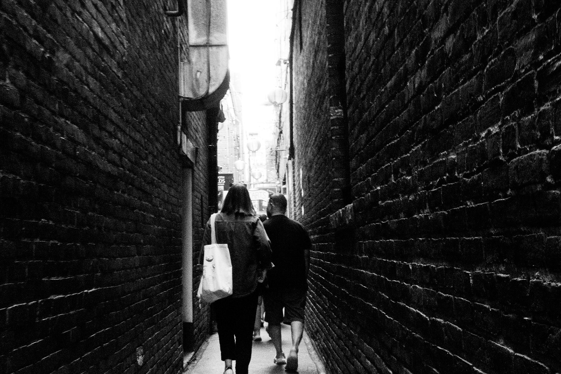 Alleyway Lovers: A monochrome photograph taken from behind a couple as they walk away from the viewer through a narrow, dark brick alleyway. Their figures are silhouetted against a brilliant, blown-out light source at the end of the passage, emphasizing the texture of the brick walls and the quiet solitude of the scene..