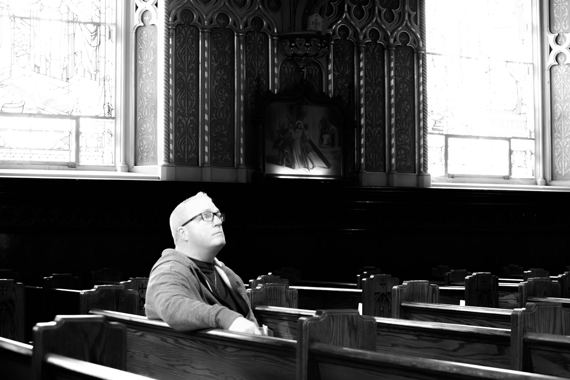 The Devotee: A monochrome photograph of a man wearing glasses, sitting alone in a wooden church pew. He is looking upward with a thoughtful, serene expression. The scene is illuminated by soft, bright light from stained-glass windows in the background, which contrasts with the dark, ornate woodwork of the church interior and the carved religious artwork behind him.