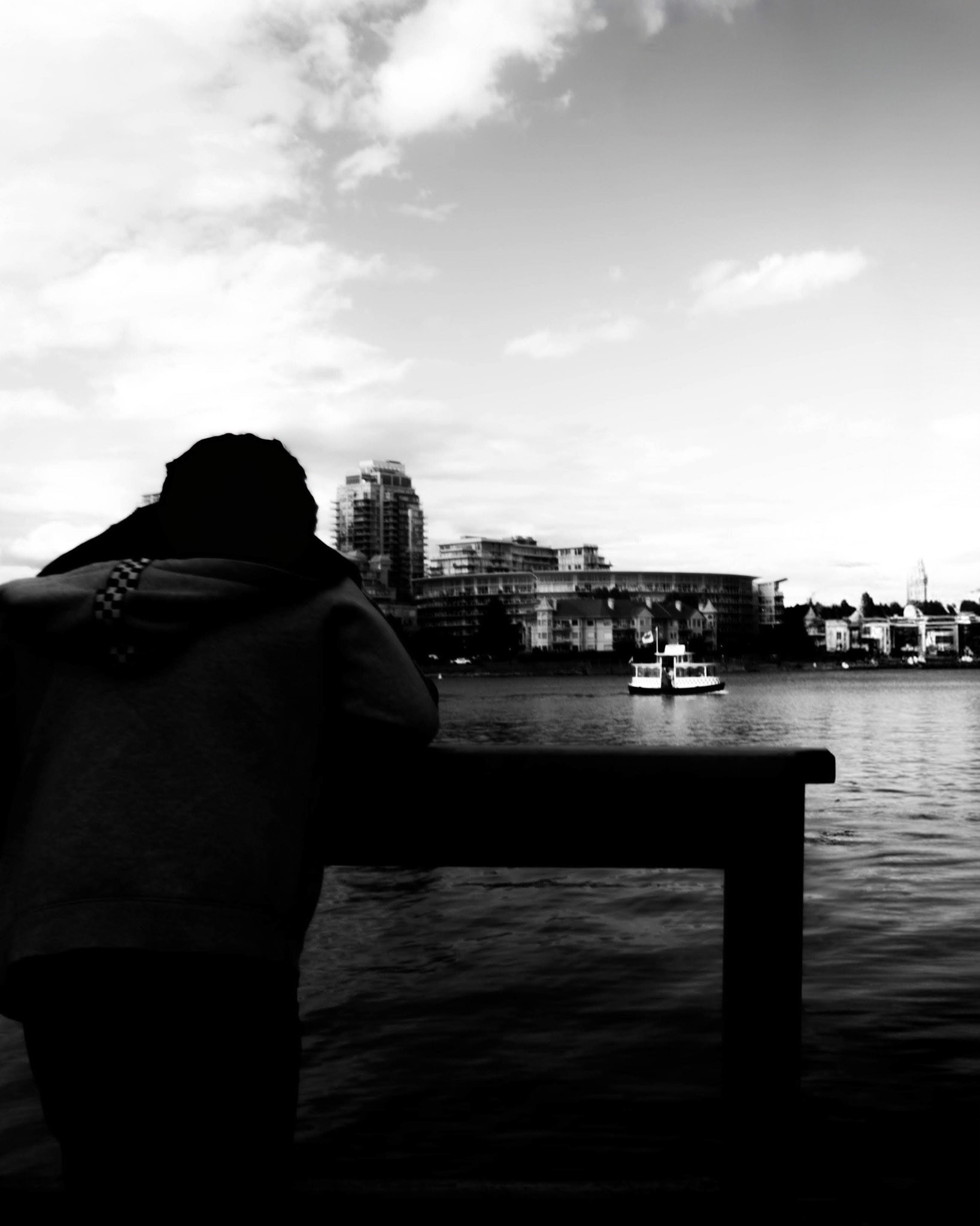 The Gazer: A monochrome photograph from a low vantage point. In the foreground, a person is shown in dark silhouette, leaning against a railing and gazing out across a body of water. In the distance, a small ferry boat travels toward a cityscape under a bright, cloudy sky. The image emphasizes the quiet solitude of the observer against the vastness of the water and urban horizon.