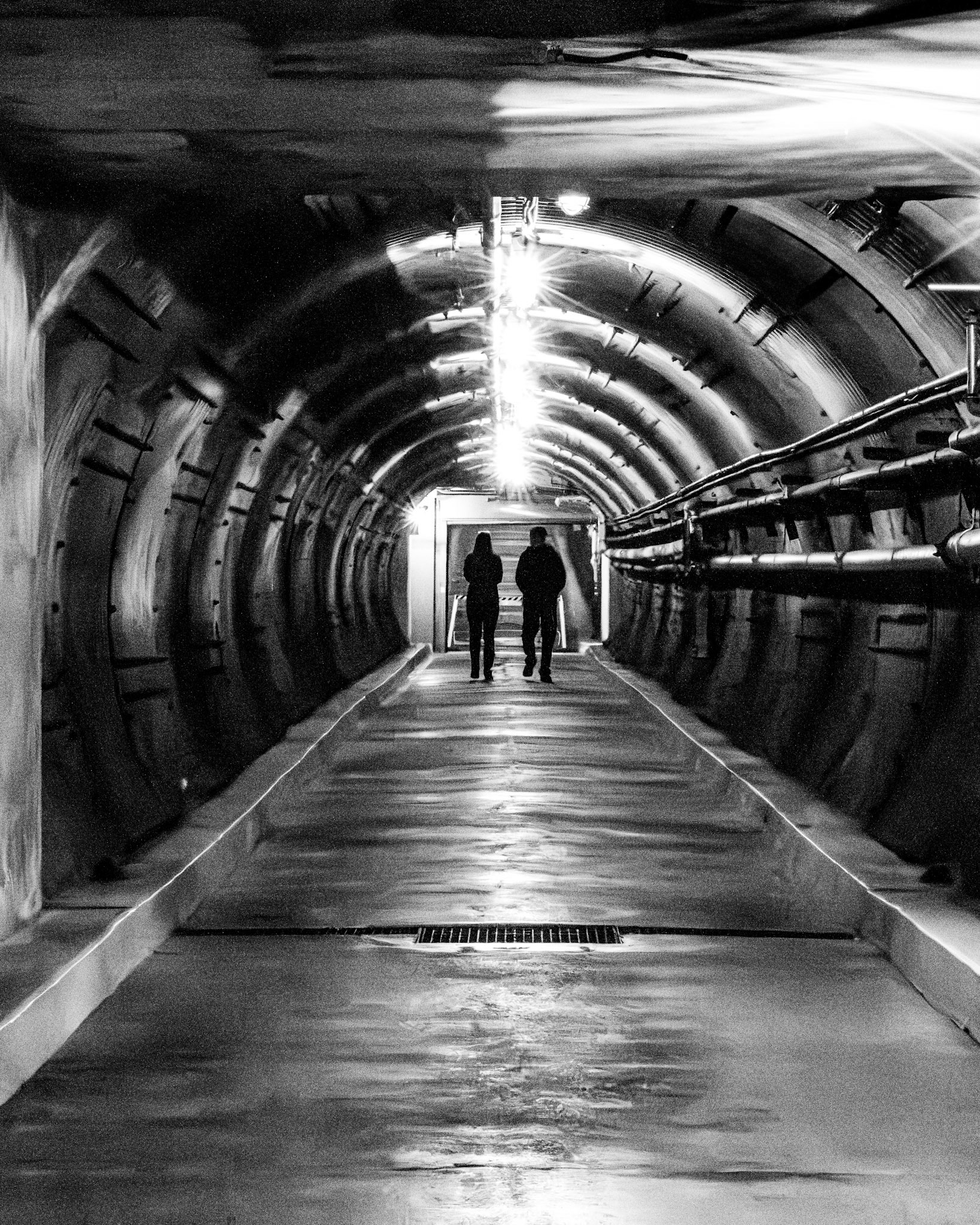 The Pilgrimage: A monochrome, symmetrical photograph looking down a long, curved industrial tunnel. Two figures are silhouetted in the distance, walking toward a bright light source at the far end of the passage. The composition is defined by the dark, textured, ribbed walls of the tunnel, overhead pipes, and strong leading lines that draw your eyes toward the subjects and the brilliant light.