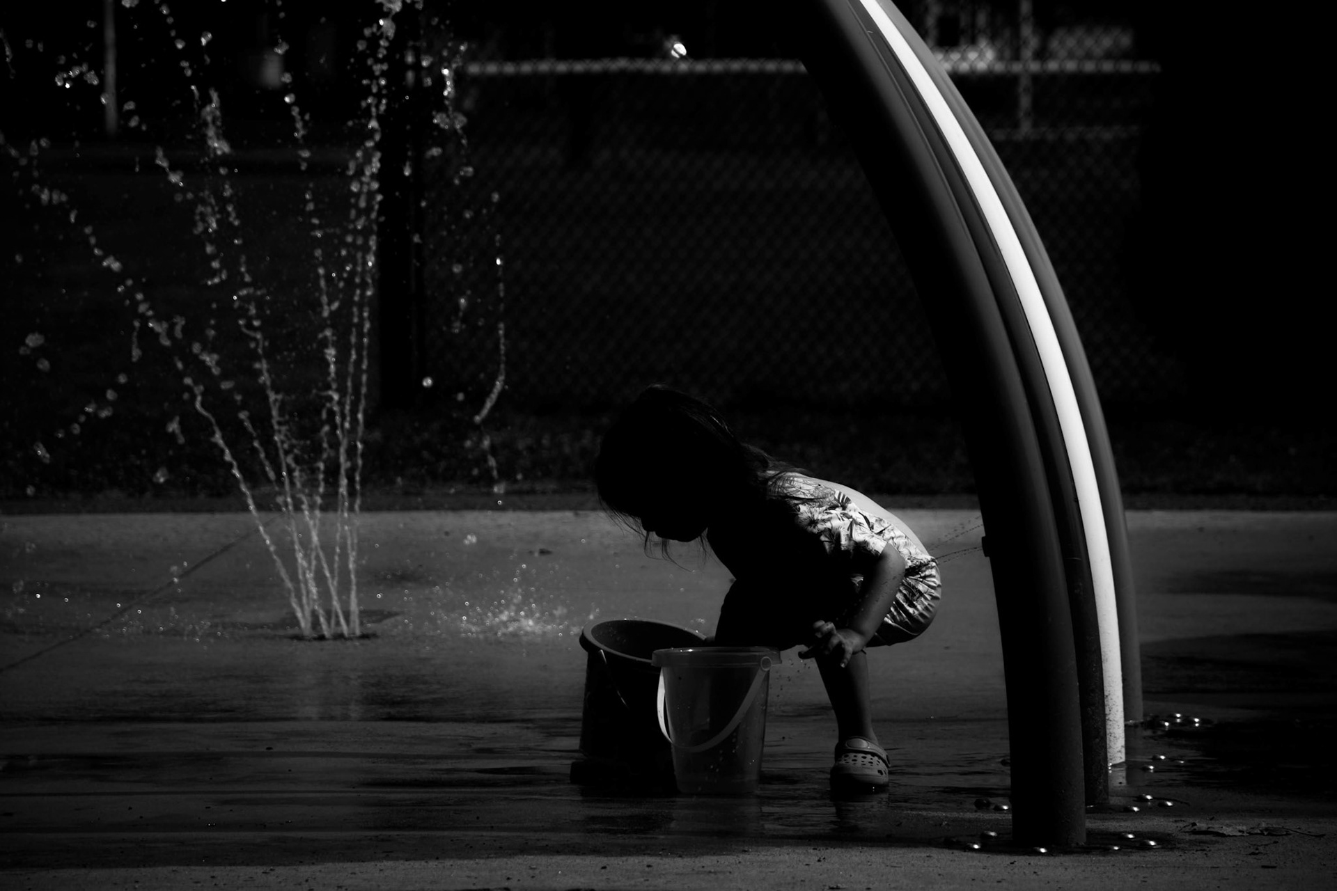 Holy Water: A monochrome photograph of a child crouching at a splash pad. The child is captured as a silhouette, intently focused on filling a plastic bucket with water from a nearby spray fountain. The scene is framed by a curved metal structure, and the contrast between the dark shadows and the sparkling water droplets creates a serene, ritualistic atmosphere.