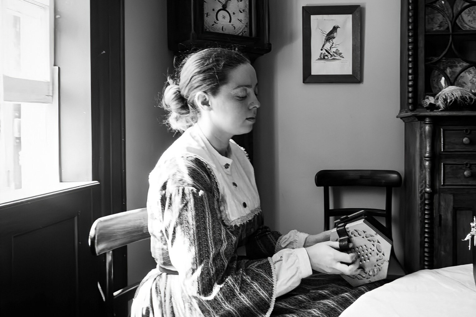 Hymn: A monochrome interior shot of a woman seated in profile near a bright window. She is dressed in period-accurate, rustic clothing and is focused intently on playing a small musical instrument, her concertina. The soft, directional light highlights her gentle expression and the texture of her dress, set against a classic, dim interior with a wall clock and framed artwork in the background.
