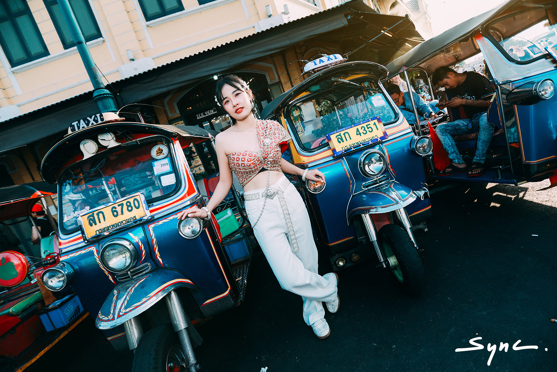 Full-body shot of a young Asian woman standing outdoors in Bangkok, wearing a striking combination of a traditional Thai sabai and casual denim jeans with Thai TukTuk car background.
