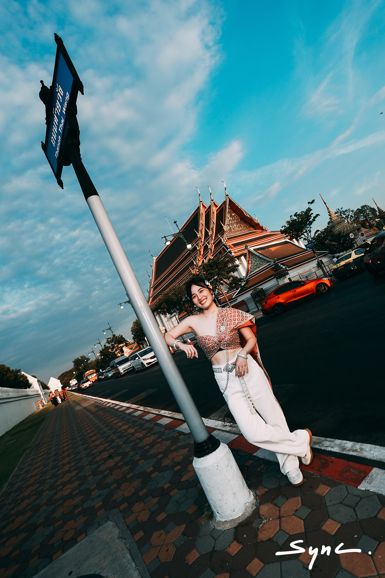Full-body shot of a young Asian woman standing outdoors in Bangkok, wearing a striking combination of a traditional Thai sabai and casual denim jeans with Thai temple background.