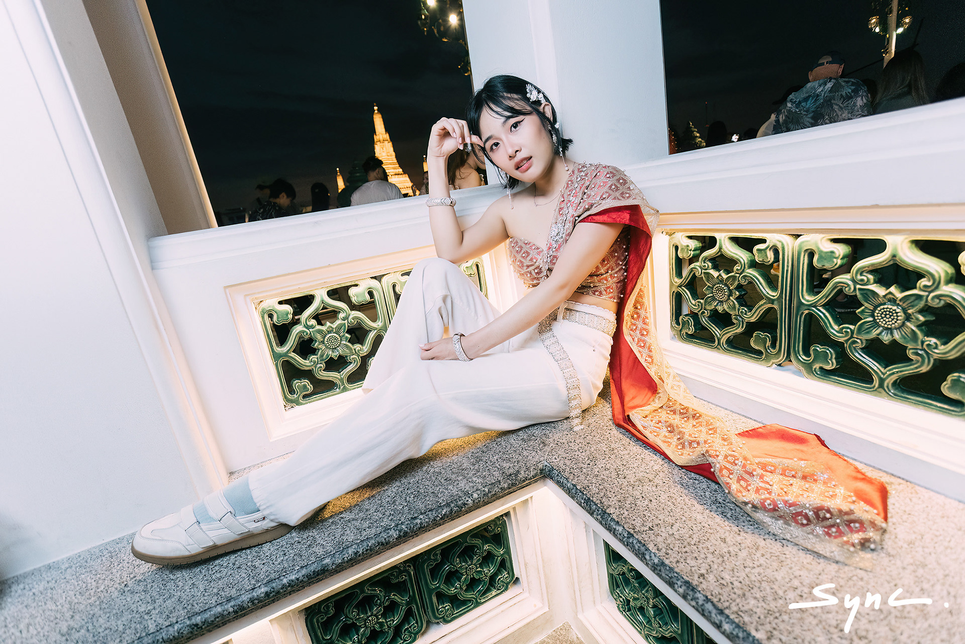 Full-body shot of a young Asian woman standing outdoors in Bangkok, wearing a striking combination of a traditional Thai sabai and casual denim jeans with Thai temple background.