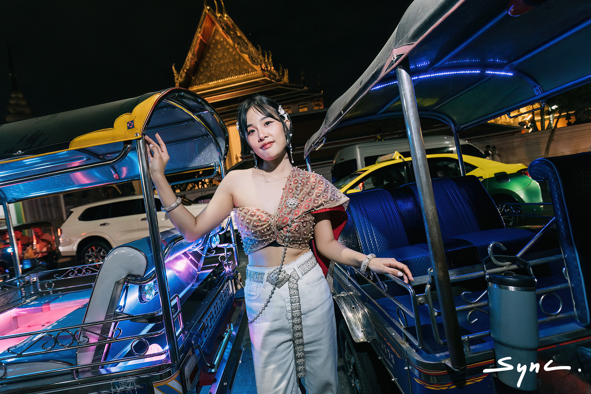 Close-up portrait of a young Asian woman modeling a fusion fashion look, wearing a traditional Thai sabai paired with denim jeans outdoors with Thai TukTuk background at night.