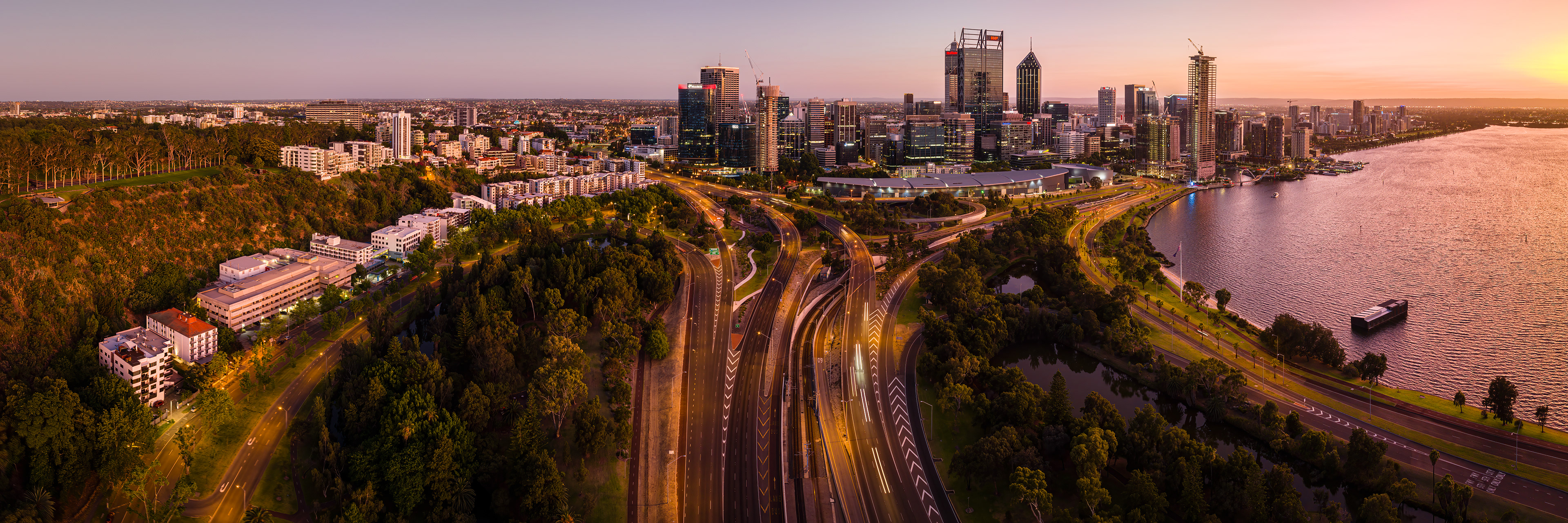 Drone panorama of Perth City with views of Kwinana Freeway entering the city, Kings Park to the left and the Swan River to the right.