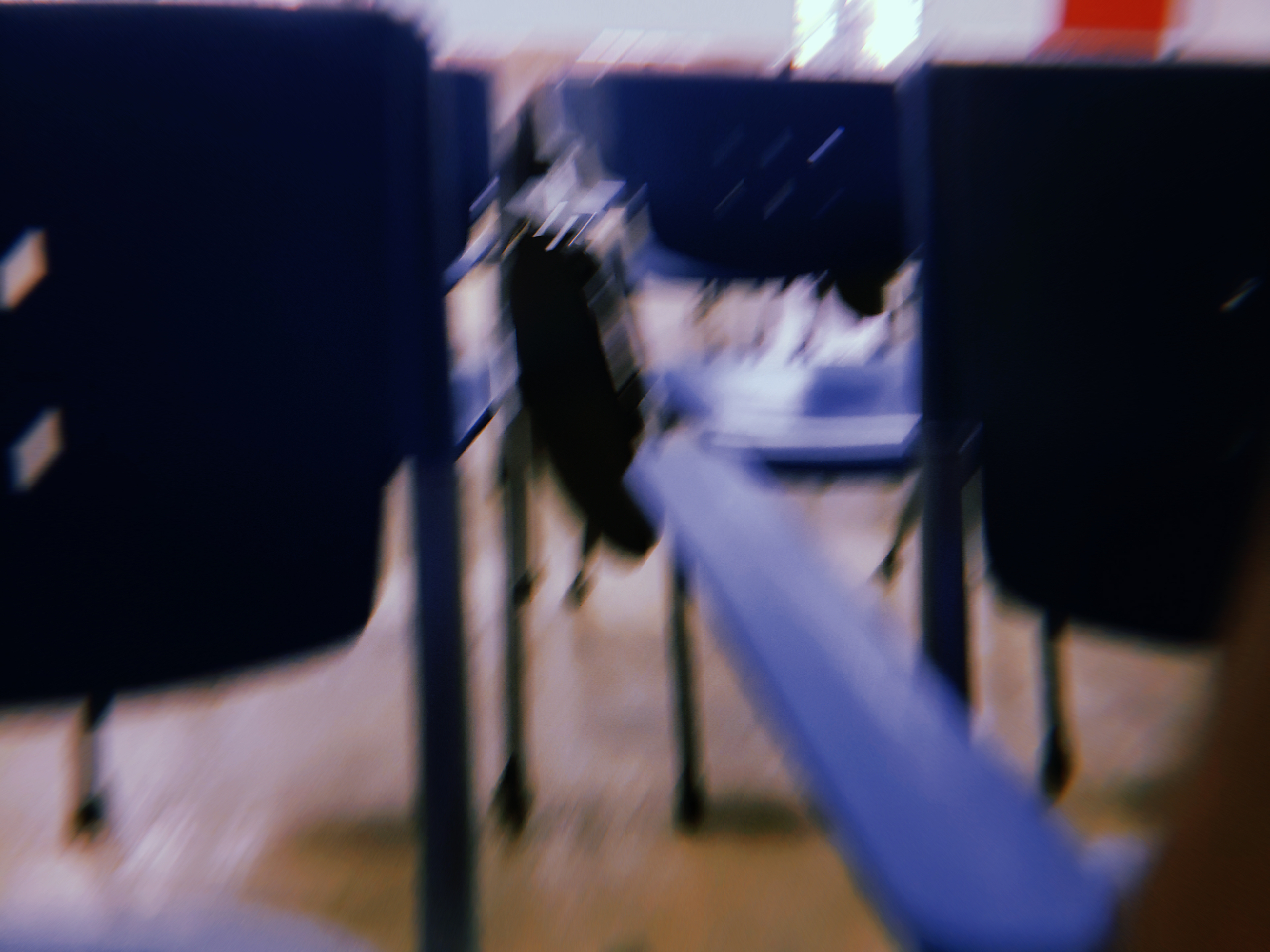 A blurred photo showing rows of blue plastic chairs, likely in a classroom or hall setting. The image is out of focus, but a black bag hanging from one of the chairs is visible in the centre. The floor appears to be tiled, and there is bright light coming from the background.