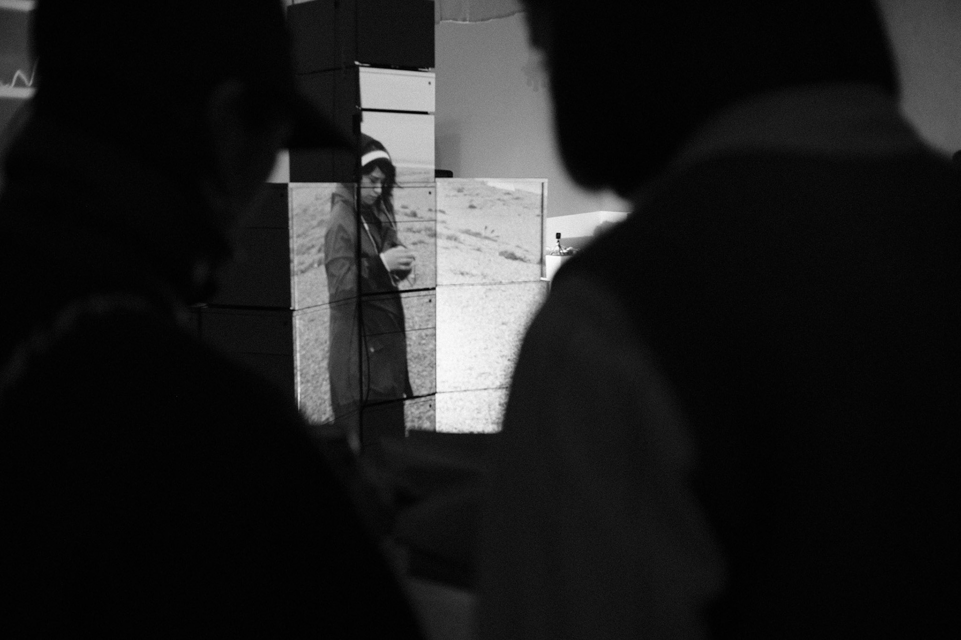 A black and white photograph taken from behind two out-of-focus figures in the foreground. They are looking at an art installation consisting of a tall, vertical stack of filing cabinets. Projected onto the structure is a video  of a woman in a coat standing in a desolate, hilly landscape.