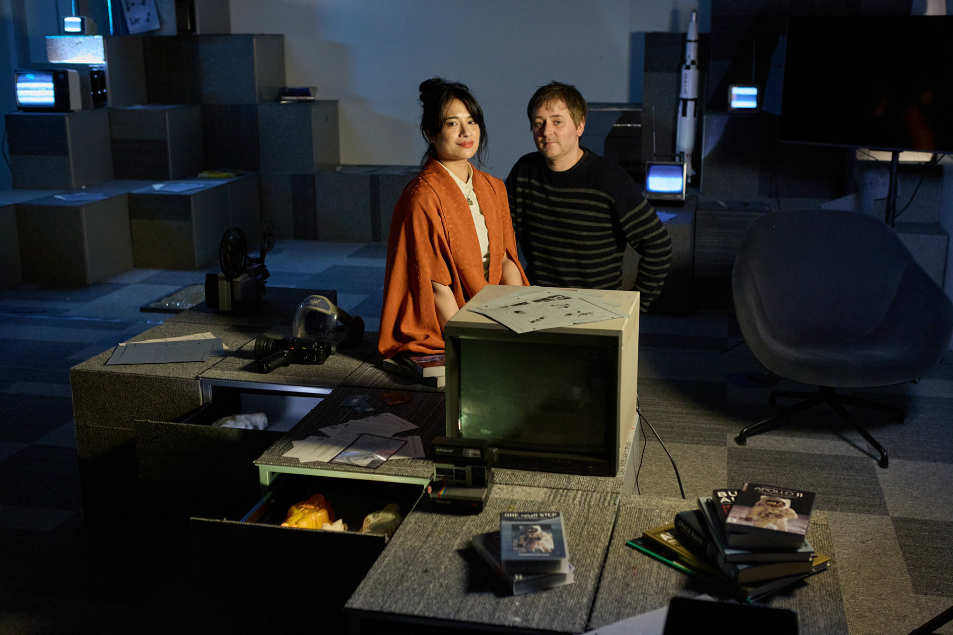 Two people sit in a dimly lit, retro-futuristic room filled with vintage technology, books, and monitors. One wears an orange shawl and looks at the camera, while the other stands behind, smiling. The space features stacked grey blocks, a rocket model, and CRT screens showing static, creating a nostalgic, sci-fi atmosphere.