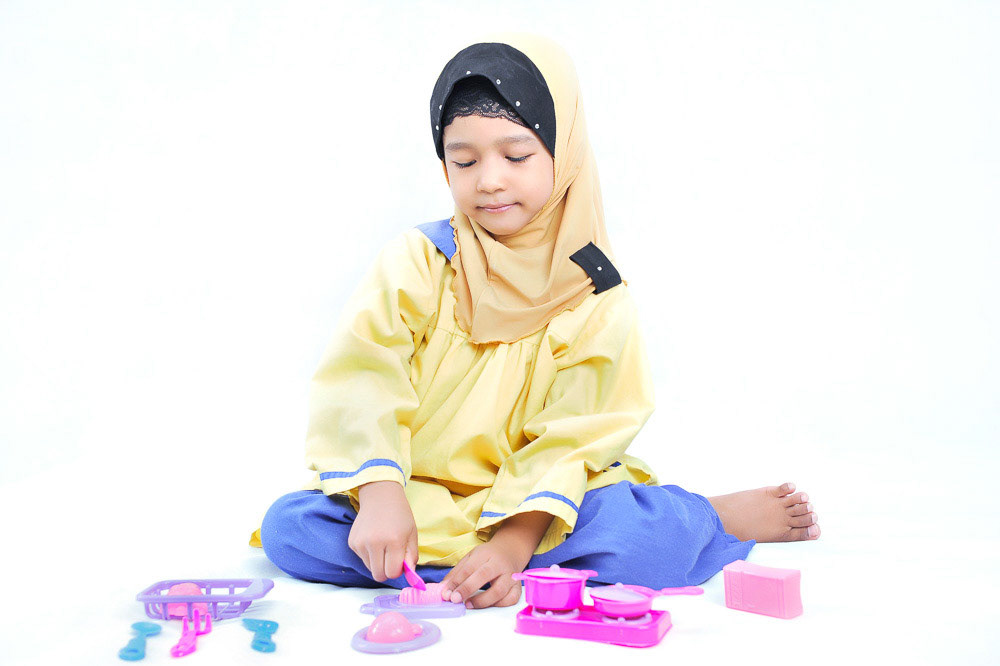 Happy, young and cute Muslim girl playing with a miniature cooking utensil toy in white isolated background.