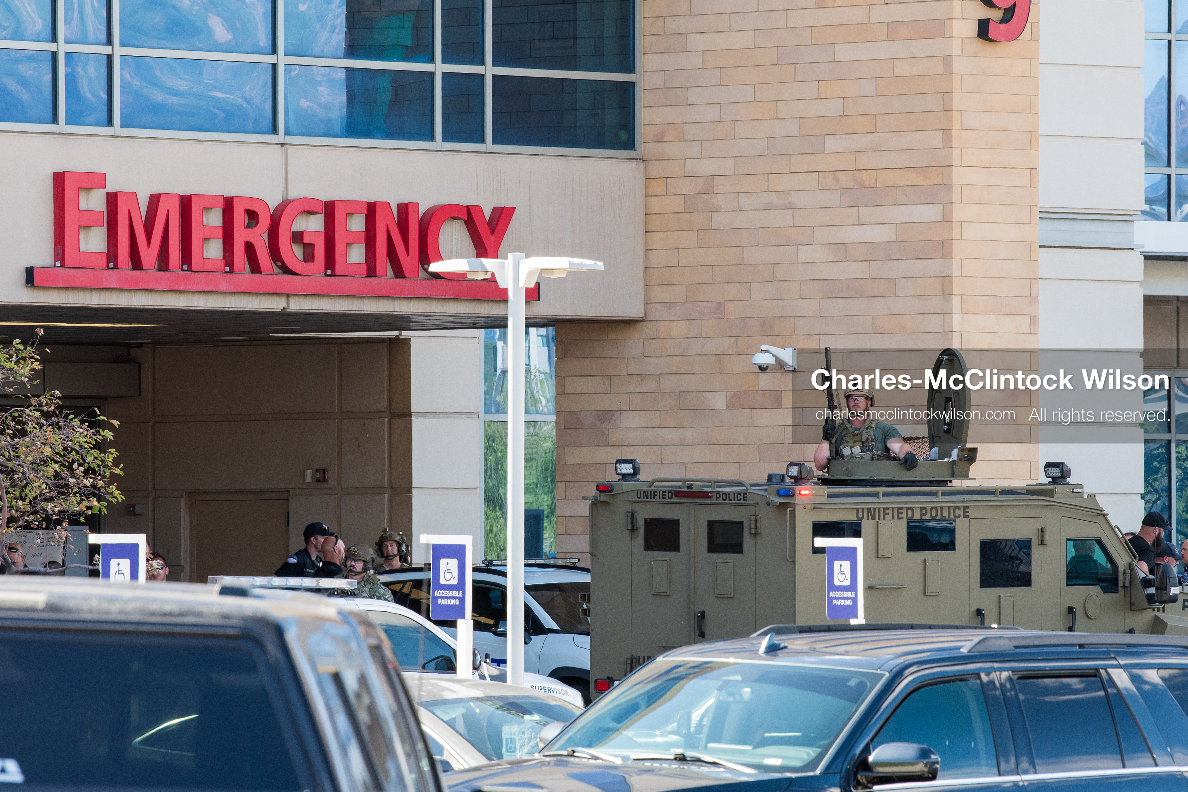 An armed law enforcement officer stands inside the roof turret hatch of an armored tactical vehicle positioned near the emergency department entrance at Intermountain Medical Center in Murray, Utah, United States, on August 17, 2026. Multiple regional police agencies secured the hospital perimeter as a safety precaution after shooting victims were admitted following a fatal shootout outside a funeral service in nearby Kearns. (Photo by Charles-McClintock Wilson/NurPhoto)