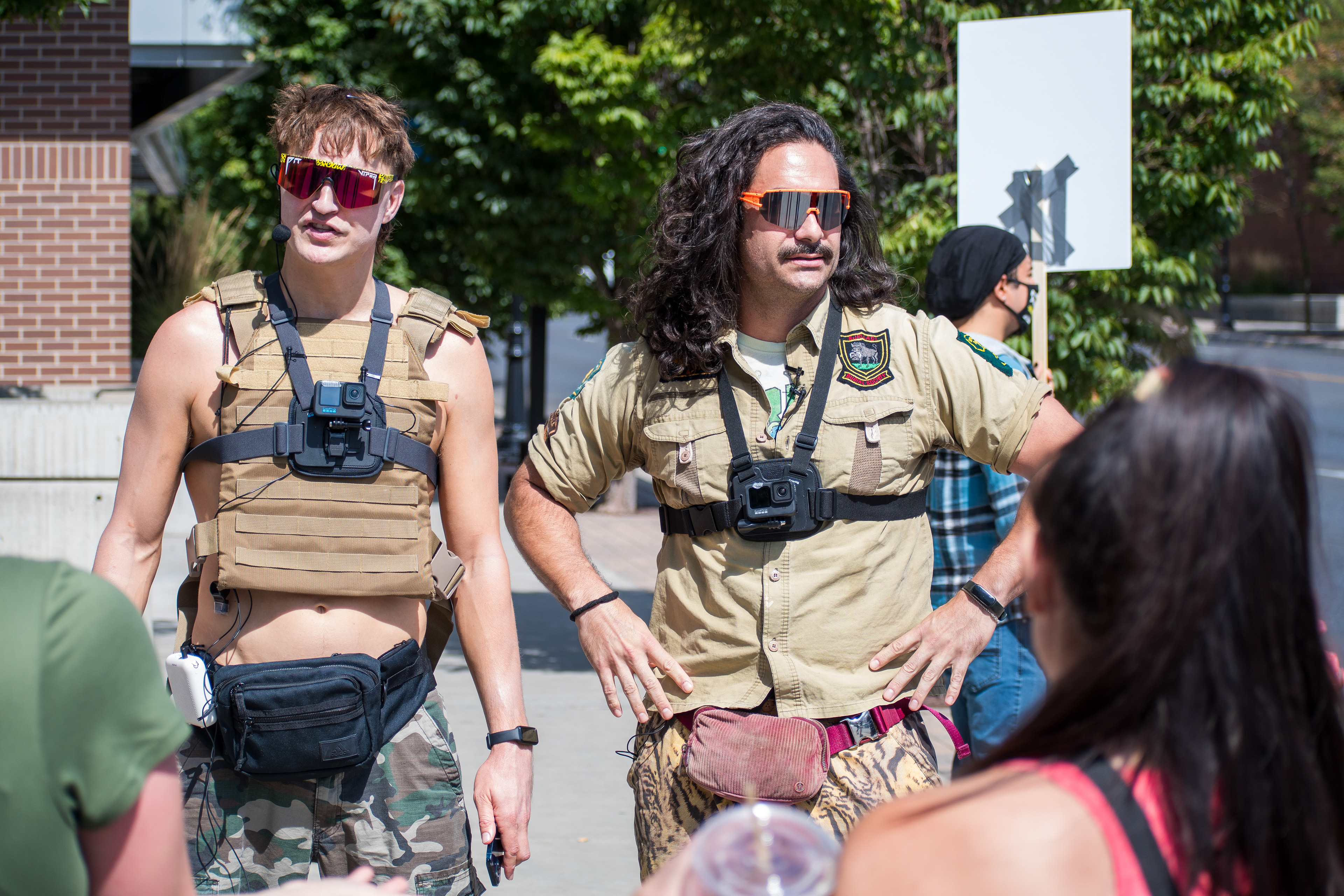 September 15, 2025 – Provo, Utah, United States: Two demonstrators in tactical-style gear stand outside the Utah Valley Convention Center during a protest against the Department of Homeland Security career expo. One wears a tan vest over a bare torso with camouflage pants and a chest-mounted camera; the other sports a tan jumpsuit with patches and a red fanny pack. Their presence adds paramilitary aesthetics to the visual landscape of civic dissent. Photograph by Charles‑McClintock Wilson / ZUMA Press Wire