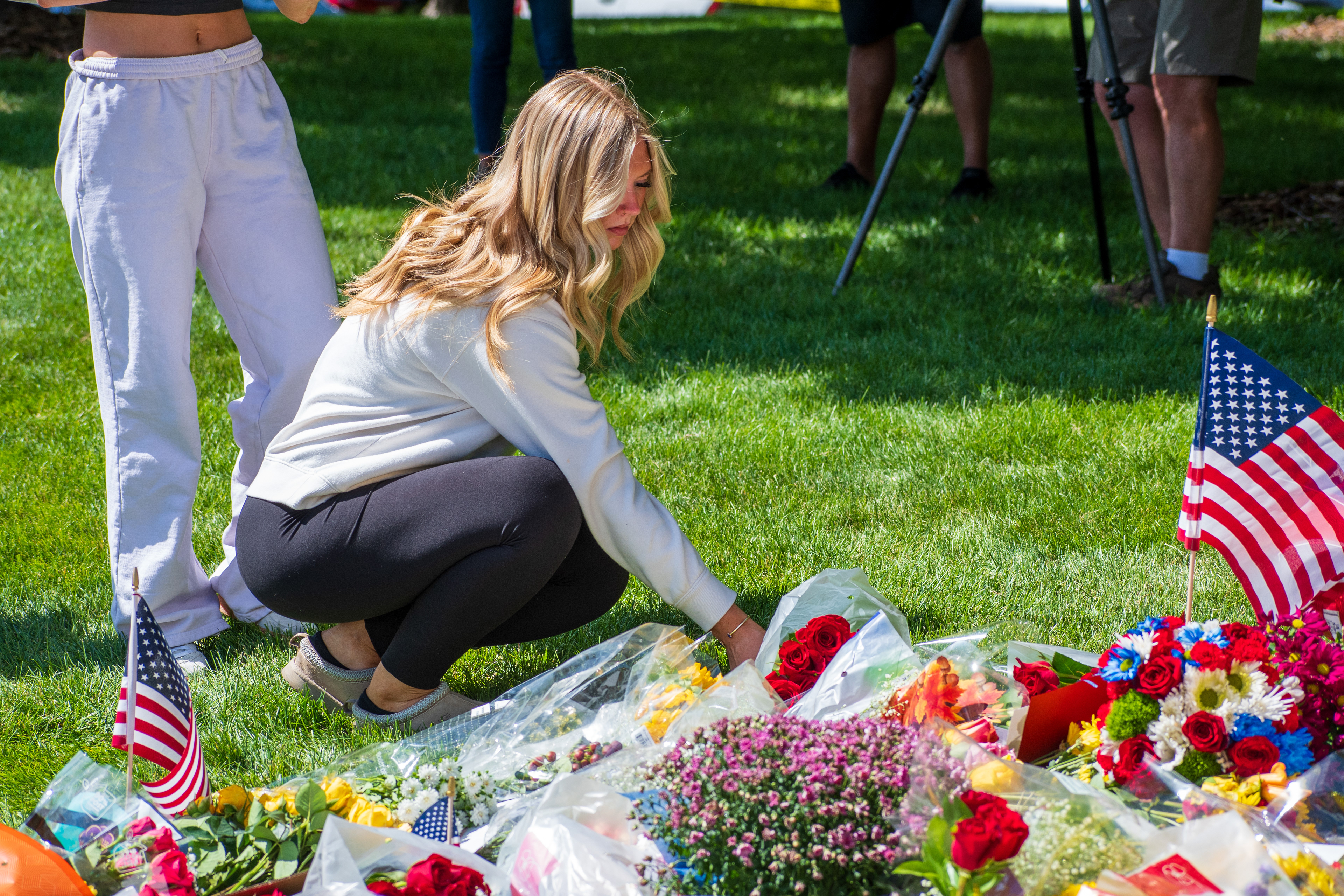 OREM, UTAH – SEPTEMBER 12, 2025: A woman kneels to place flowers at a memorial site for Charlie Kirk near Utah Valley University. Surrounded by bouquets, American flags, and fellow mourners, the tribute reflects a moment of quiet remembrance and communal grief. © Charles‑McClintock Wilson / ZUMA Press
