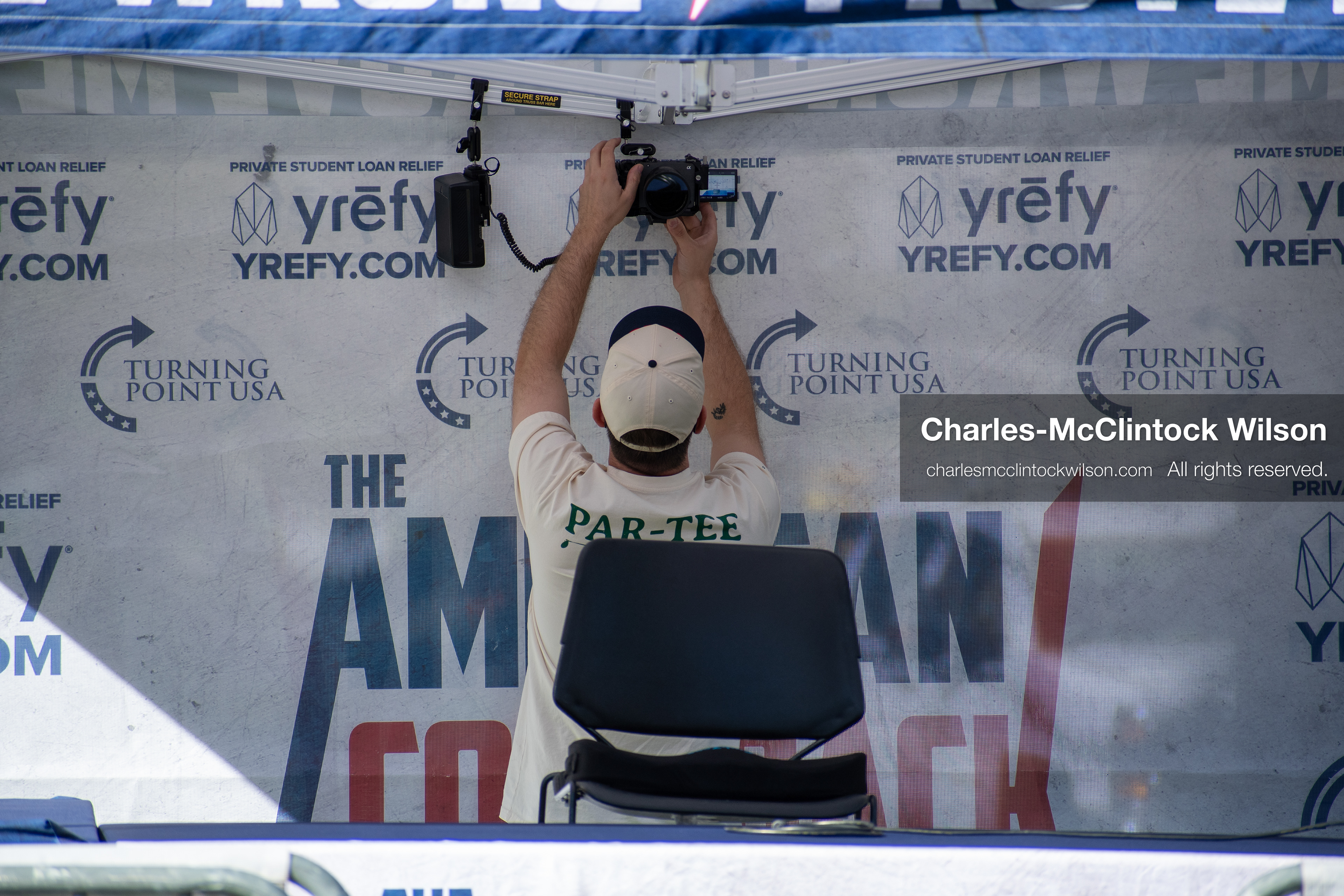 OREM, UTAH – SEPTEMBER 10, 2025: An event technician adjusts equipment inside a branded booth at Utah Valley University during the opening stop of the American Comeback Tour. Framed in mid-action beneath signage for student loan relief, the individual reflects the quiet coordination and logistical precision that shaped the event’s infrastructure. The image captures a moment of setup, branding, and operational clarity. © Charles-McClintock Wilson / ZUMA Press
