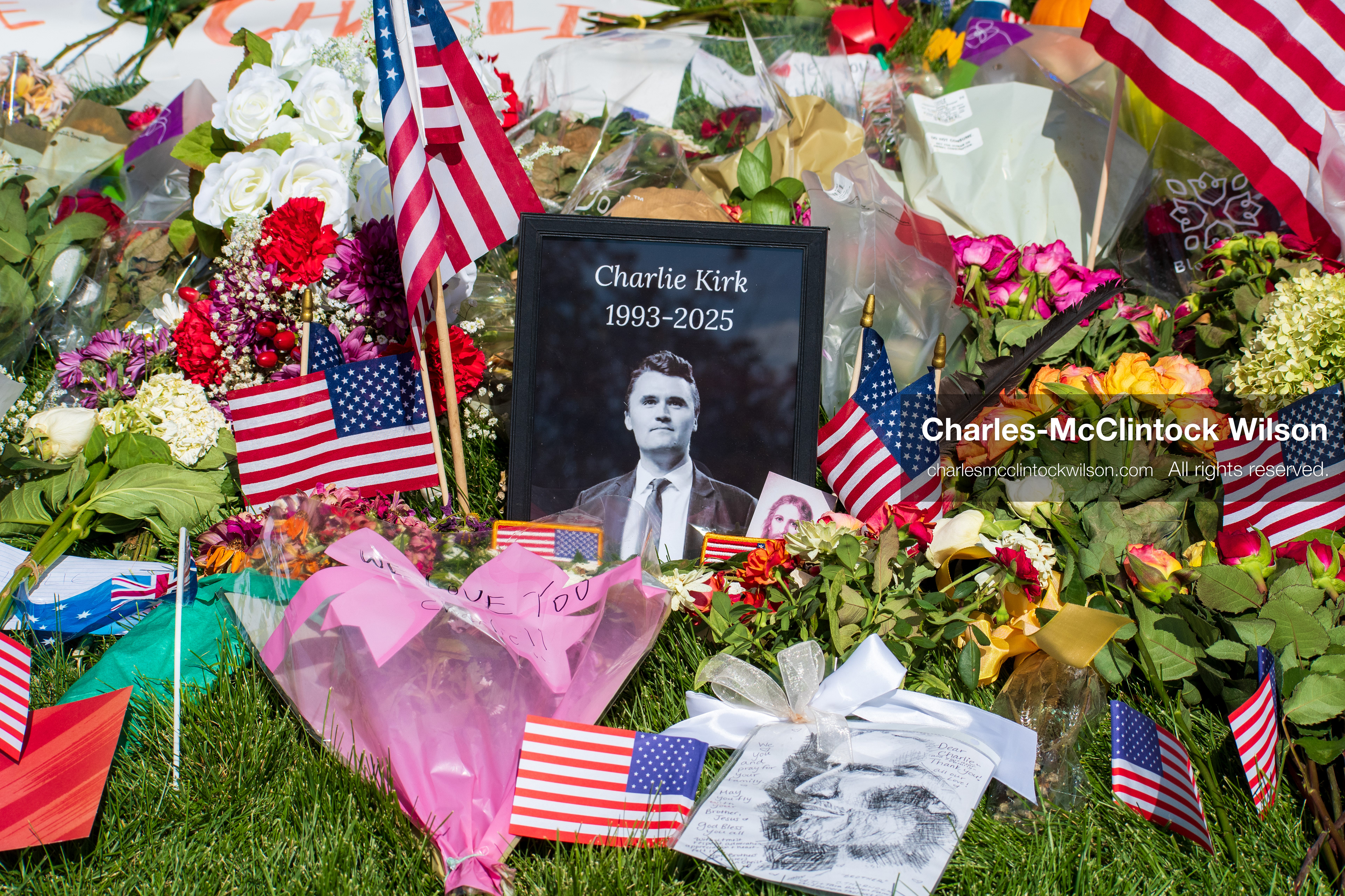 OREM, UTAH – SEPTEMBER 15, 2025: A framed photograph labeled “Charlie Kirk 1993–2025” is displayed at a memorial site on the campus of Utah Valley University. American flags, flowers, and tribute items are arranged on the grass surrounding the portrait. © Charles‑McClintock Wilson / ZUMA Press