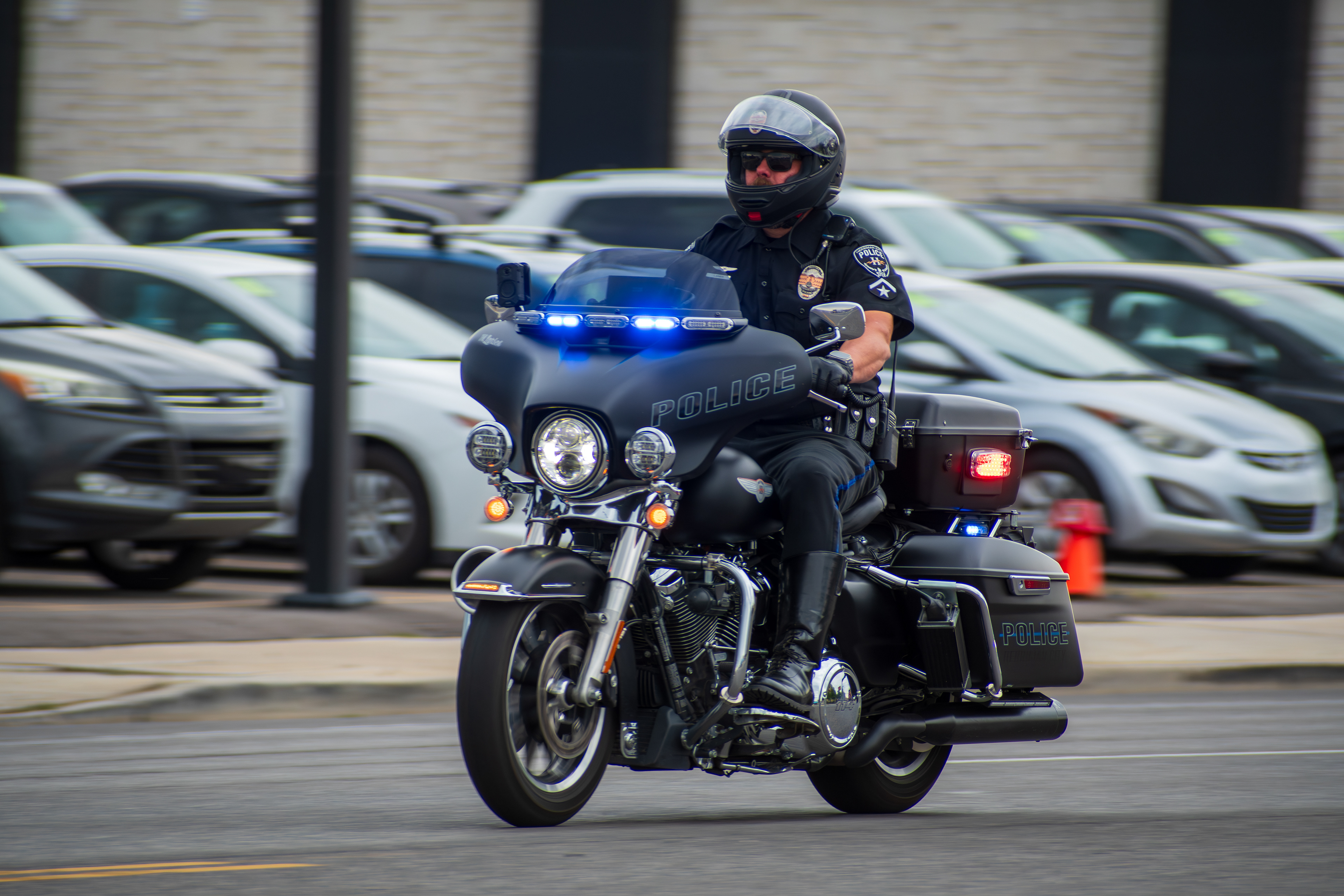 Murray, Utah — Sept. 8, 2025: A police officer navigates a city street on a black motorcycle with blue lights activated, passing a parking lot and brick-faced building.