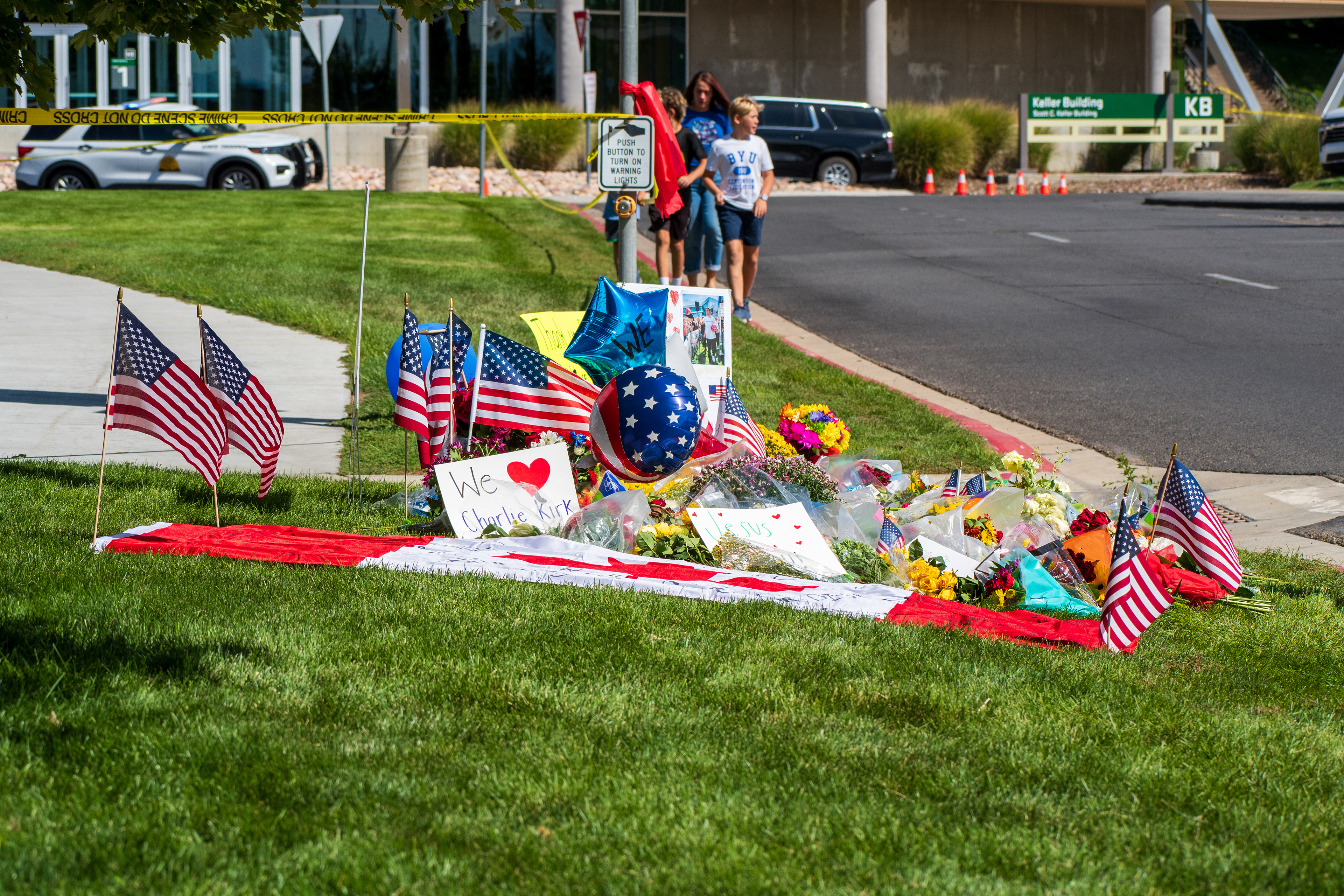 OREM, UTAH – SEPTEMBER 12, 2025: A memorial site for Charlie Kirk is seen on the lawn beside the Keller Building at Utah Valley University. American flags, flowers, balloons, and handwritten posters surround a red-and-white cloth laid out among the tributes. Two adults and two children walk toward the display. © Charles‑McClintock Wilson / ZUMA Press