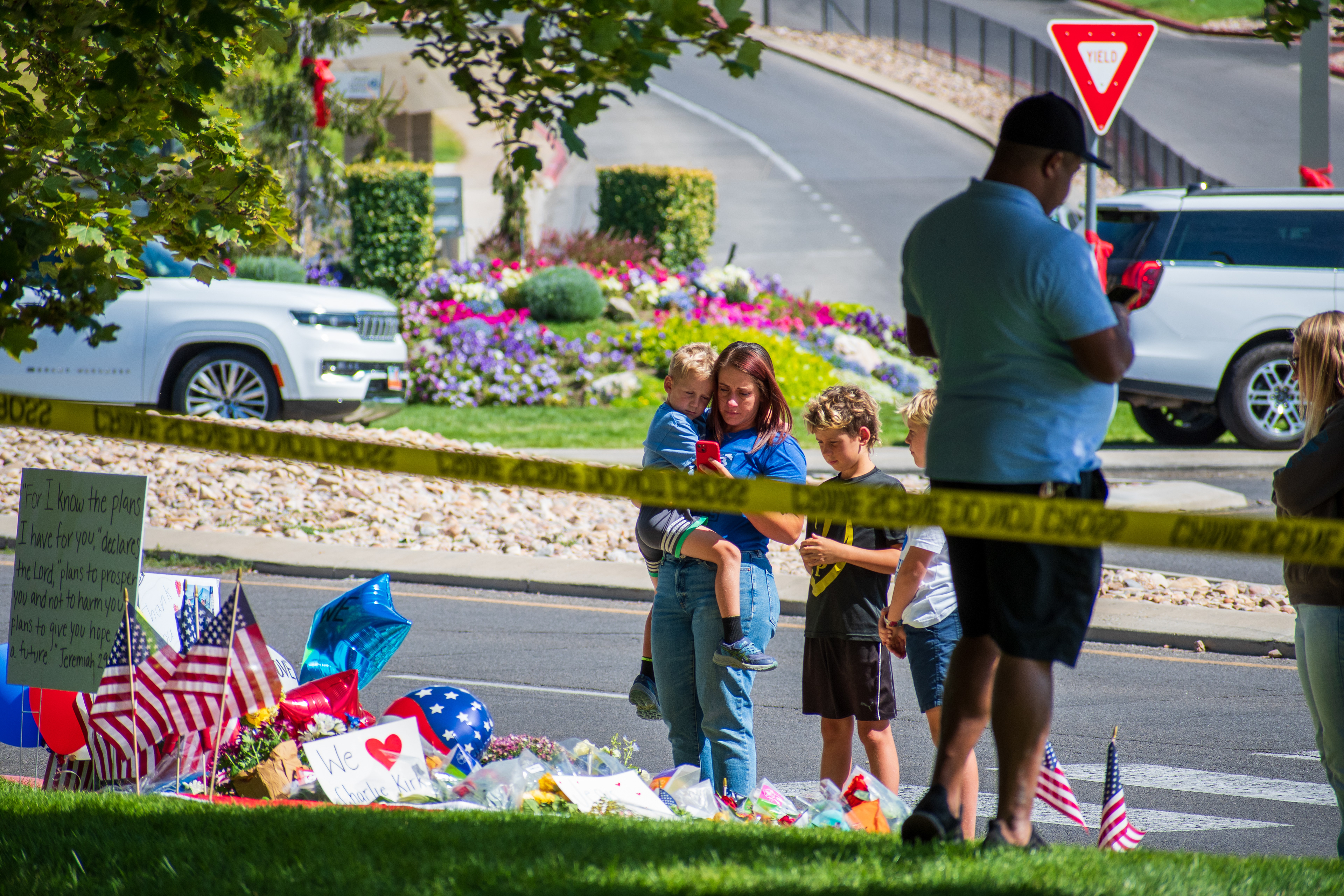 OREM, UTAH – SEPTEMBER 12, 2025: A woman holding a child stands beside a young boy and a man at a memorial site for Charlie Kirk near Utah Valley University. The tribute includes American flags, balloons, flowers, and handwritten posters arranged along the roadside. © Charles‑McClintock Wilson / ZUMA Press