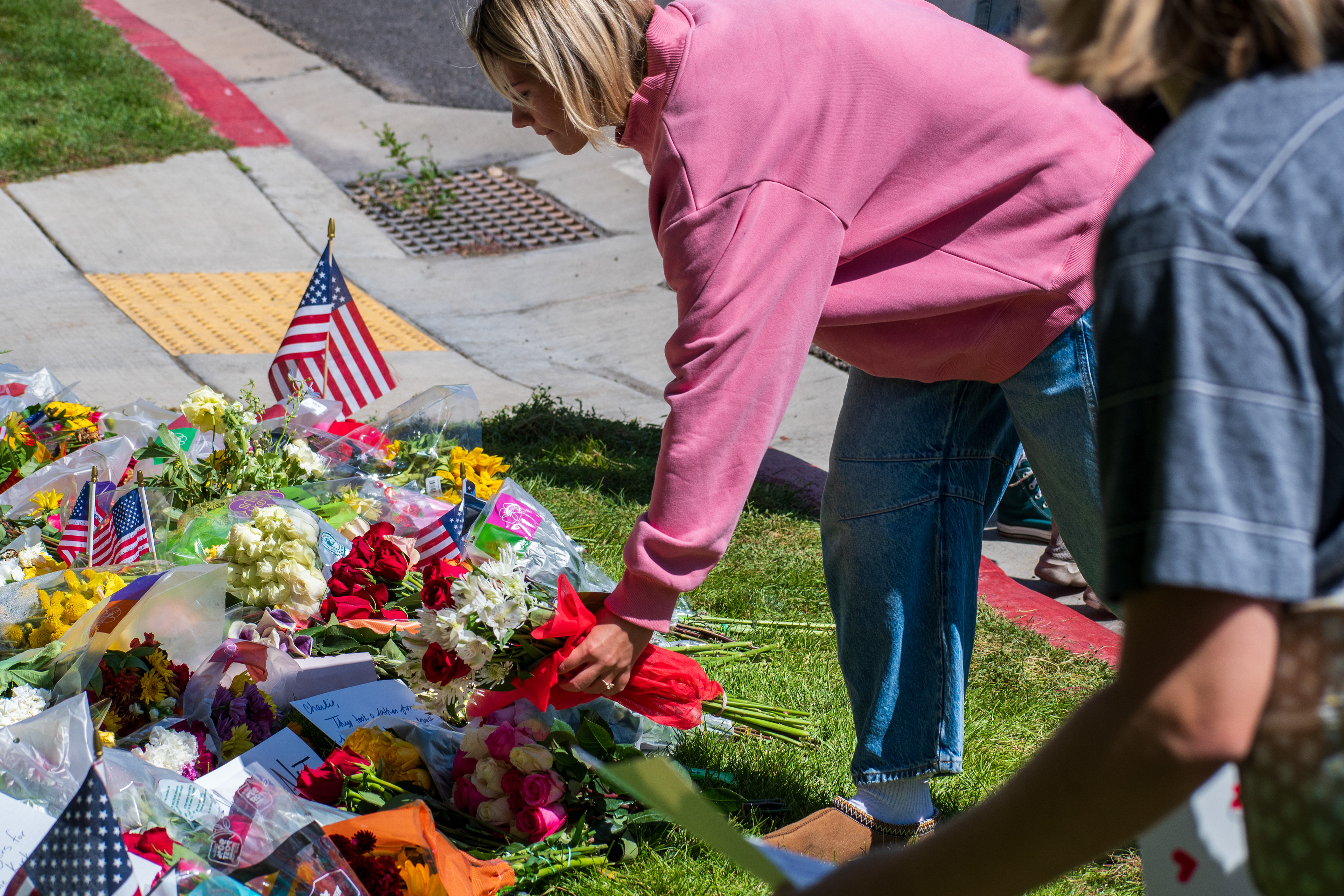 OREM, UTAH – SEPTEMBER 12, 2025: A woman in a pink hoodie adjusts flowers at a memorial site for Charlie Kirk near Utah Valley University. Bouquets, small American flags, and handwritten tributes are arranged on the grass beside the sidewalk as mourners gather. © Charles‑McClintock Wilson / ZUMA Press 