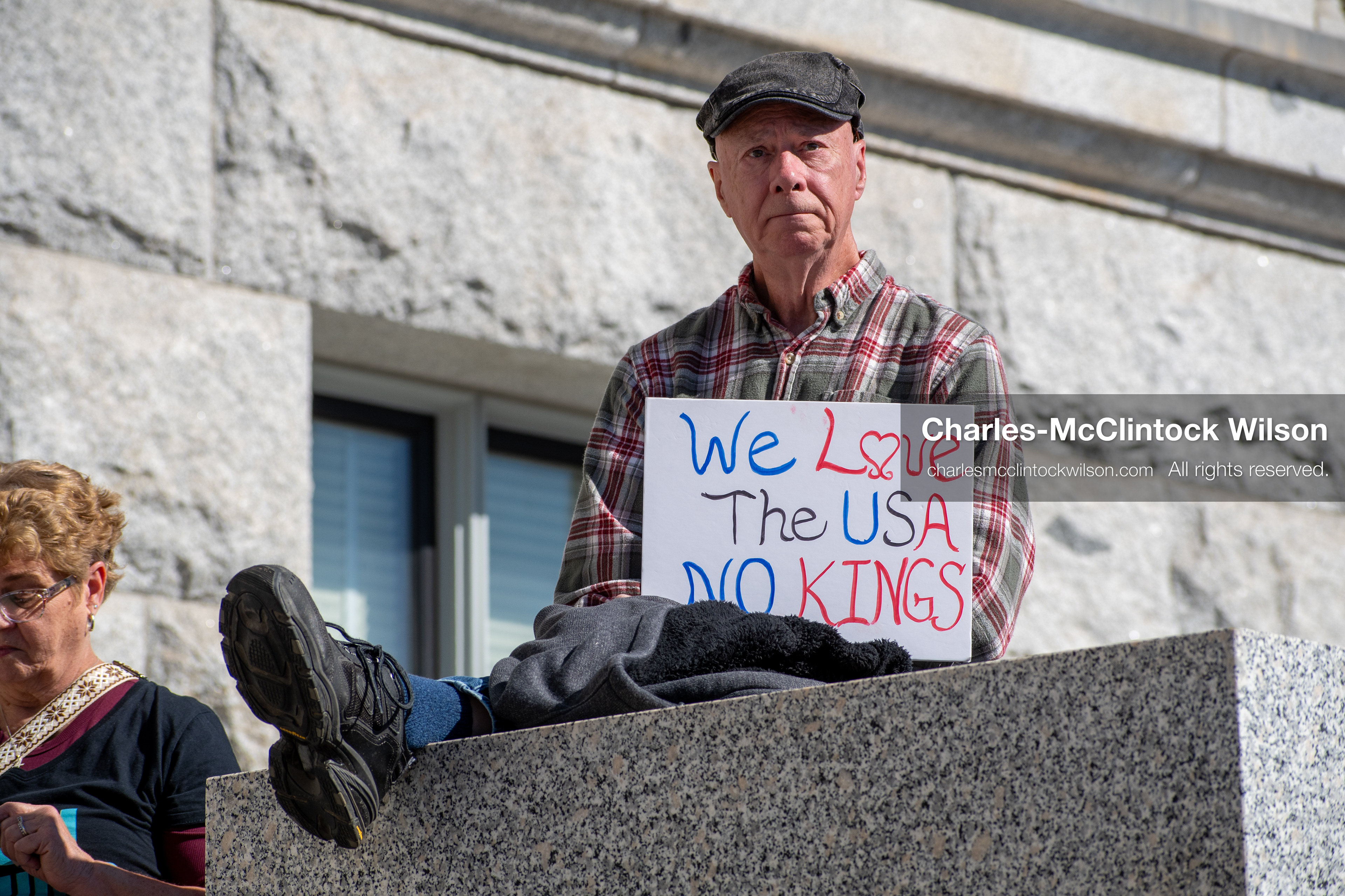 October 18, 2025, Salt Lake City, Utah, USA: A demonstrator holds a sign while seated on a stone ledge during a "No Kings" protest at the Utah State Capitol. The protest was part of a nationwide mobilization.