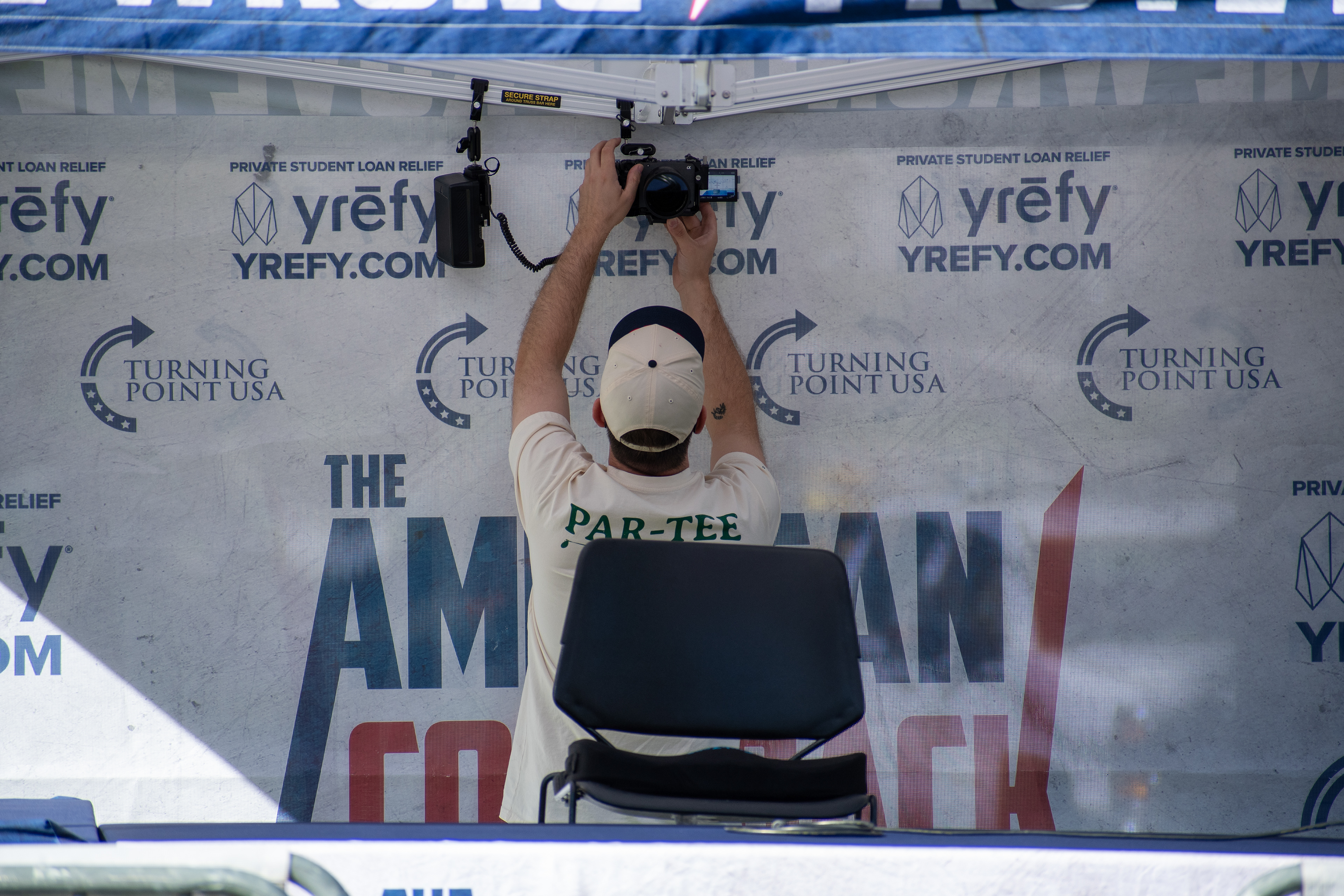 OREM, UTAH – SEPTEMBER 10, 2025: An event technician adjusts equipment inside a branded booth at Utah Valley University during the opening stop of the American Comeback Tour. Framed in mid-action beneath signage for student loan relief, the individual reflects the quiet coordination and logistical precision that shaped the event’s infrastructure. The image captures a moment of setup, branding, and operational clarity. © Charles-McClintock Wilson / ZUMA Press