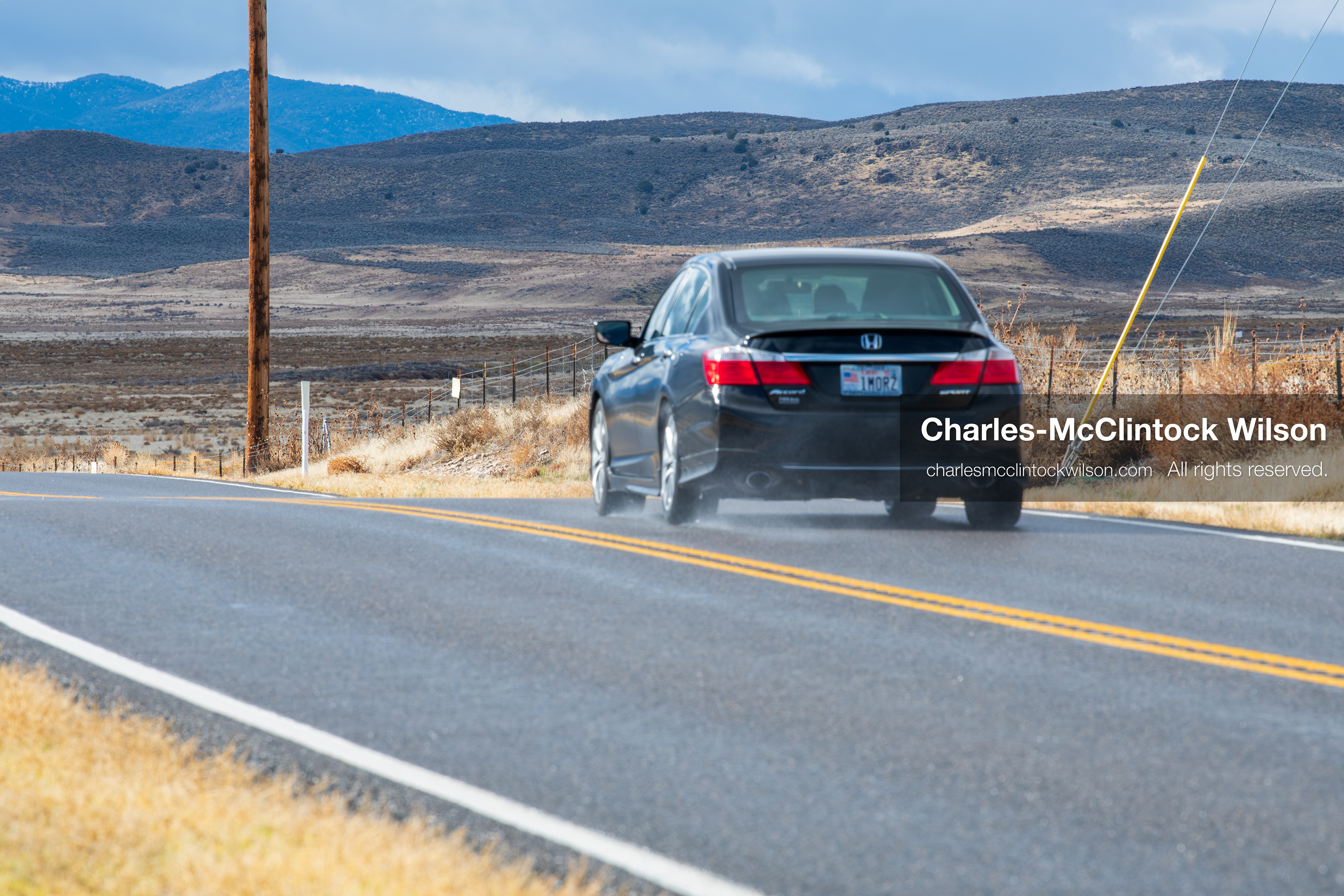 January 1, 2026, Saratoga Springs, Utah, USA: A black sedan travels along a rural road near Utah Lake on January 1, 2026, in Saratoga Springs, Utah, USA. Tire spray reflects wet conditions as dry grass, utility poles, and distant mountains frame the landscape under partly cloudy skies. (Credit Image: © Charles-McClintock Wilson/ZUMA Press Wire)