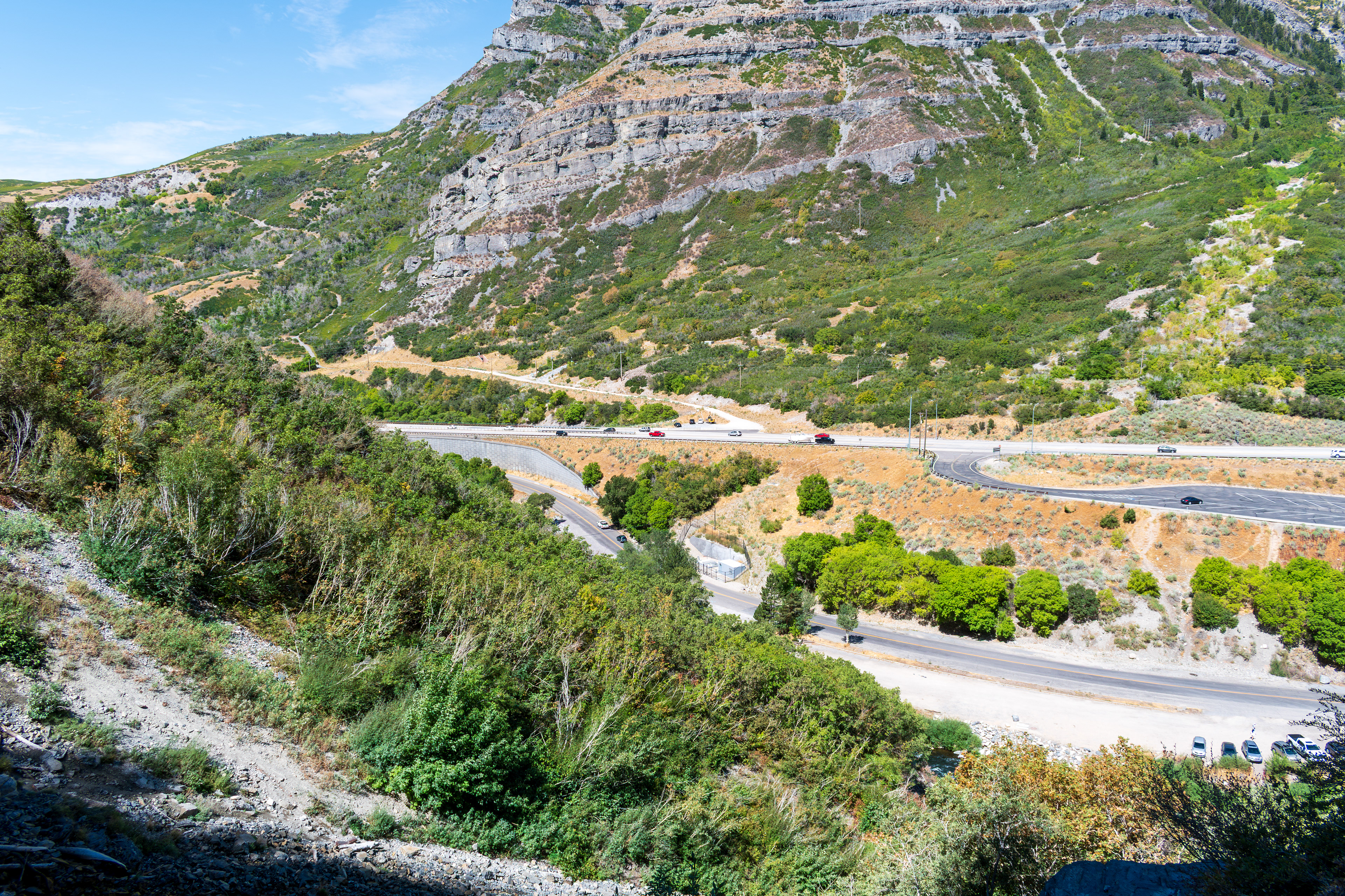 Provo, Utah, US — Sept 1, 2025: A scenic view from Bridal Veil Falls captures steep, layered rock formations and lush vegetation, with vehicles winding through the rugged terrain along U.S. Route 189.