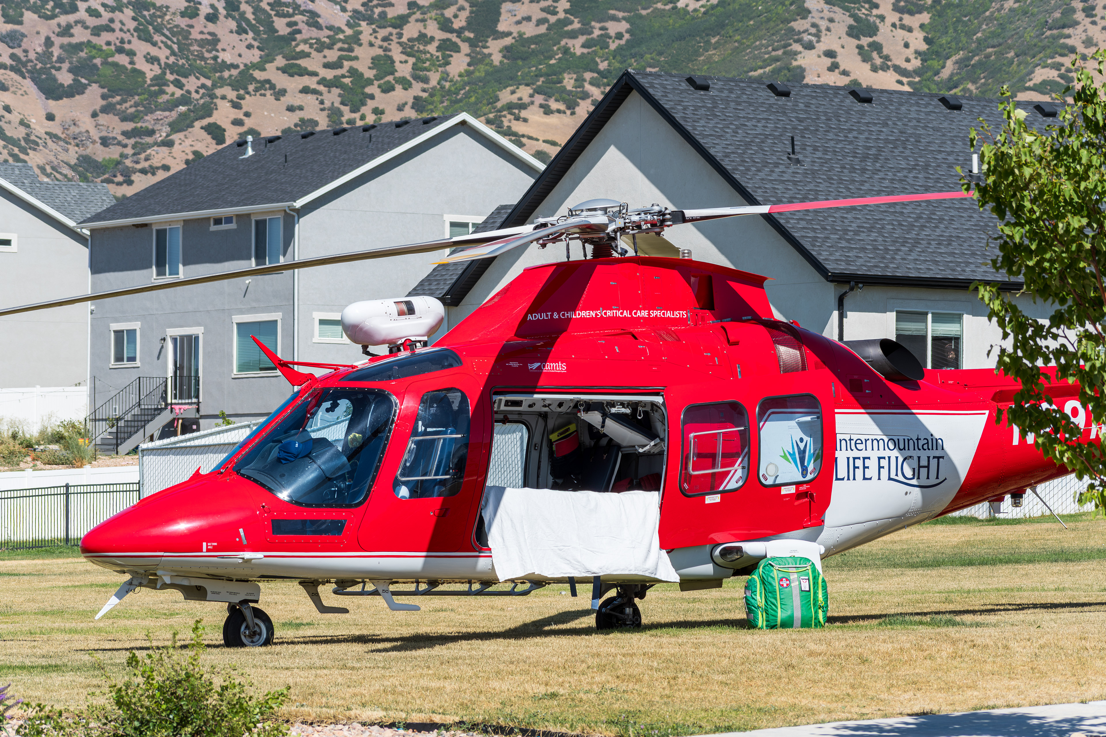  Santaquin, Utah, USA — September 1, 2025: A red Intermountain Life Flight medical helicopter is parked on a grassy area near residential homes, with a green tarp visible in the foreground. 