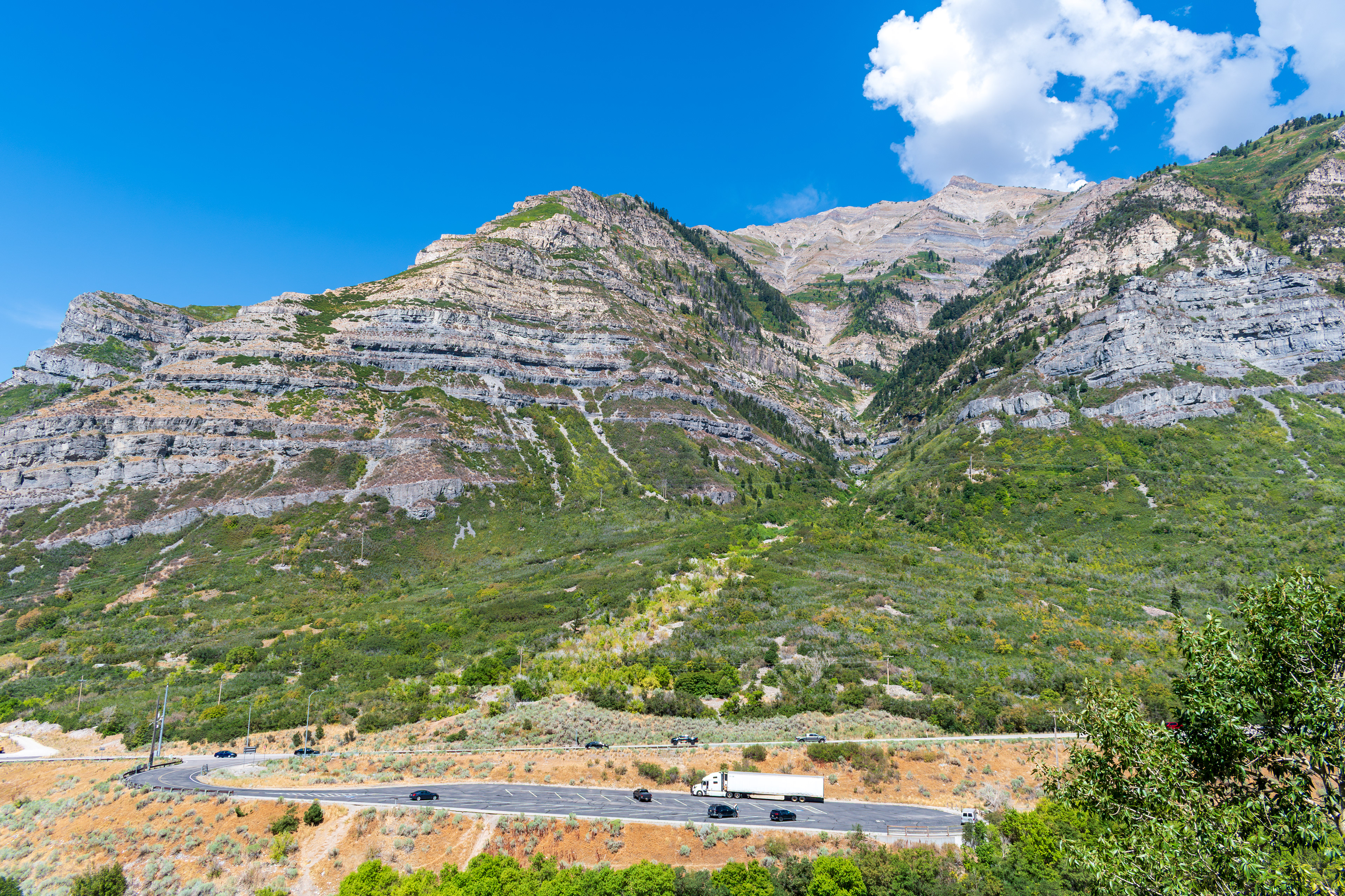 Provo, Utah, US — Sept 1, 2025: A scenic view from Bridal Veil Falls captures steep, layered rock formations and lush vegetation, with vehicles winding through the rugged terrain along U.S. Route 189.