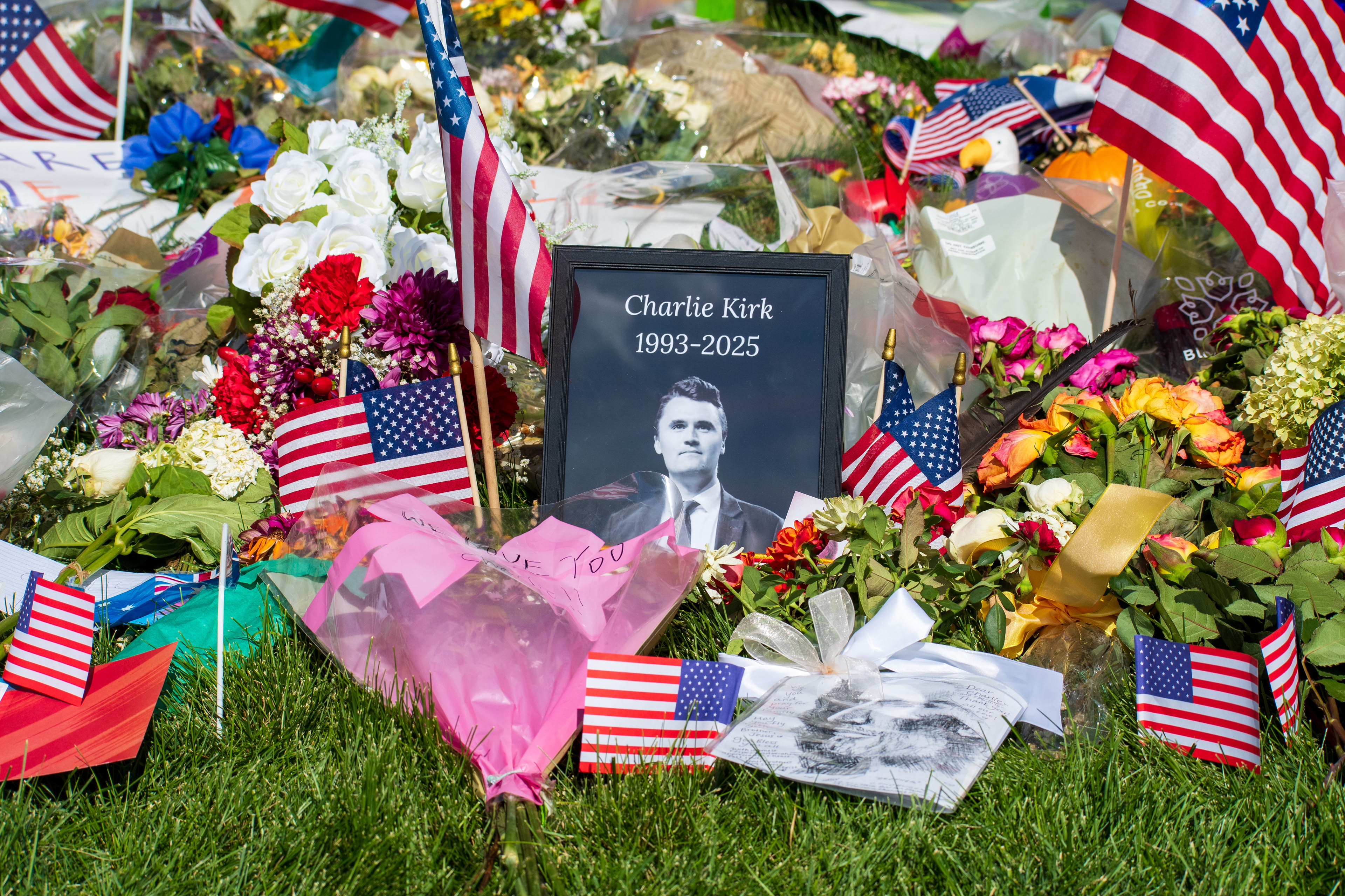OREM, UTAH – SEPTEMBER 15, 2025: A framed photograph labeled “Charlie Kirk 1993–2025” is displayed at a memorial site on the campus of Utah Valley University. American flags, flowers, and tribute items are arranged on the grass surrounding the portrait. © Charles‑McClintock Wilson / ZUMA Press